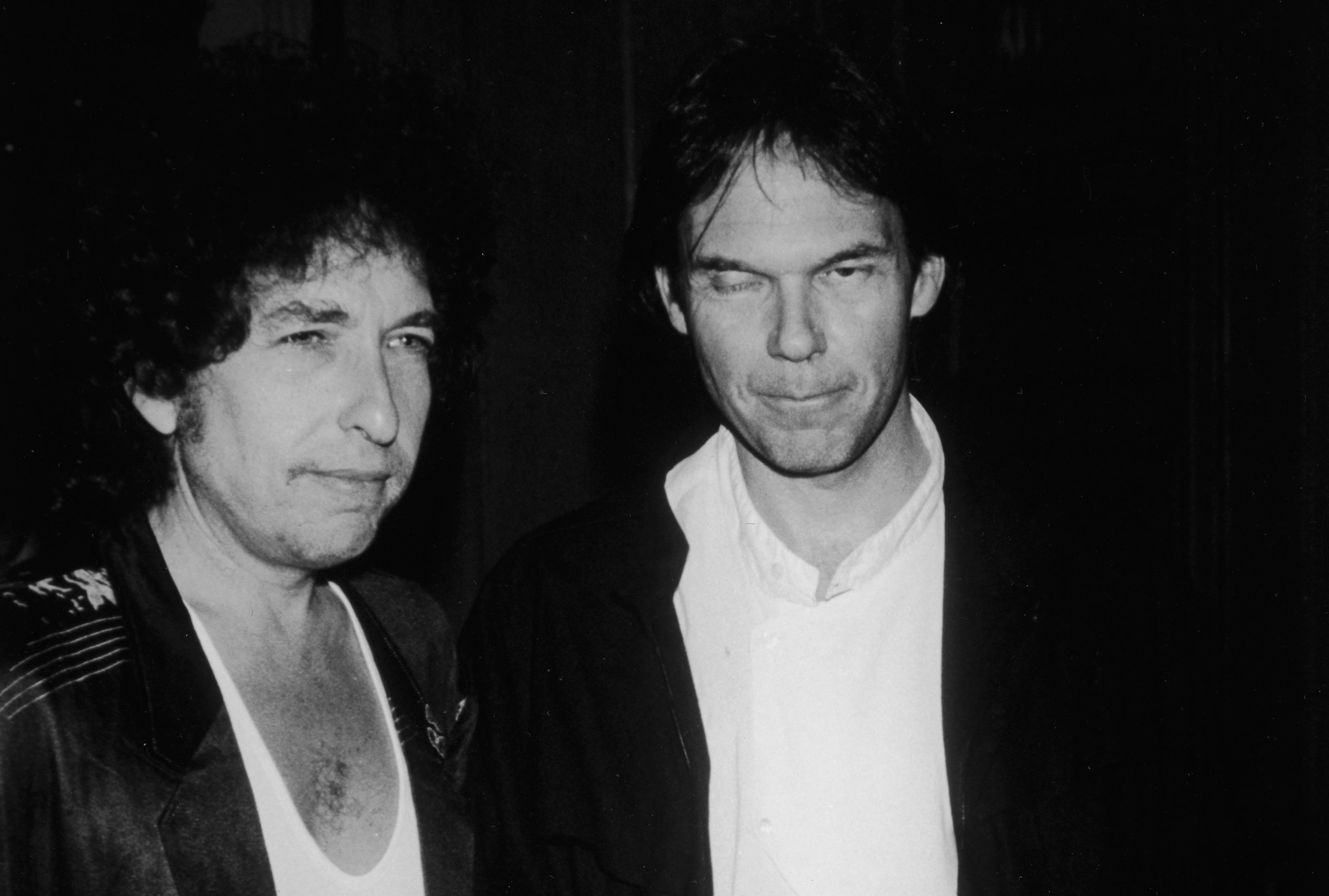 A black and white photo of Bob Dylan and Neil Young both wearing white shirts and black jackets.