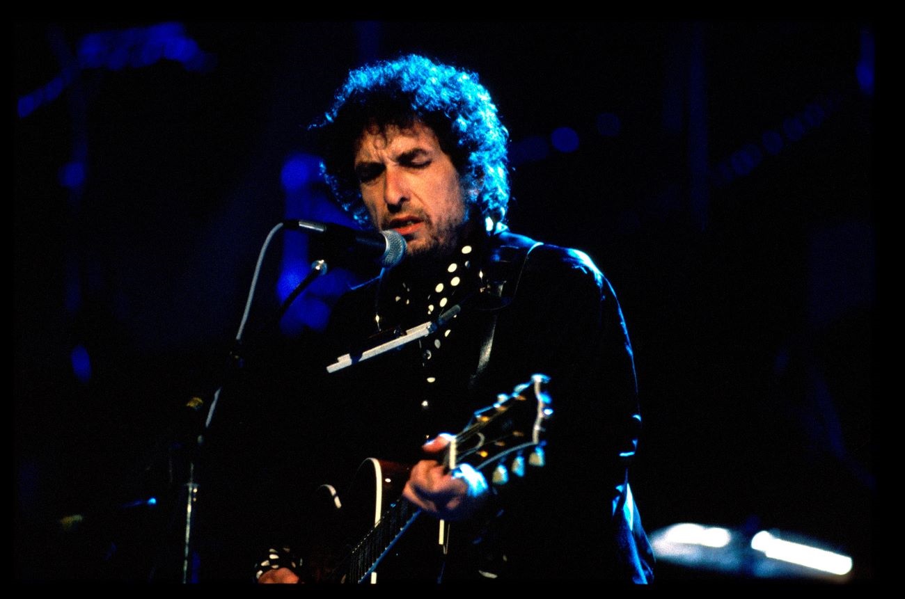 Bob Dylan plays guitar and sings into a microphone during a show in 1991.