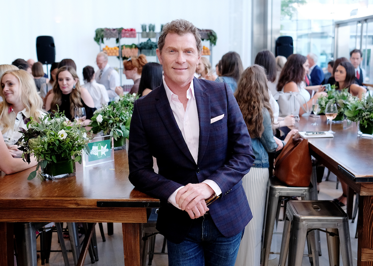 Bobby Flay, who has a Bulgur Salad recipe, smiles as he leans against a table wearing a blue suit