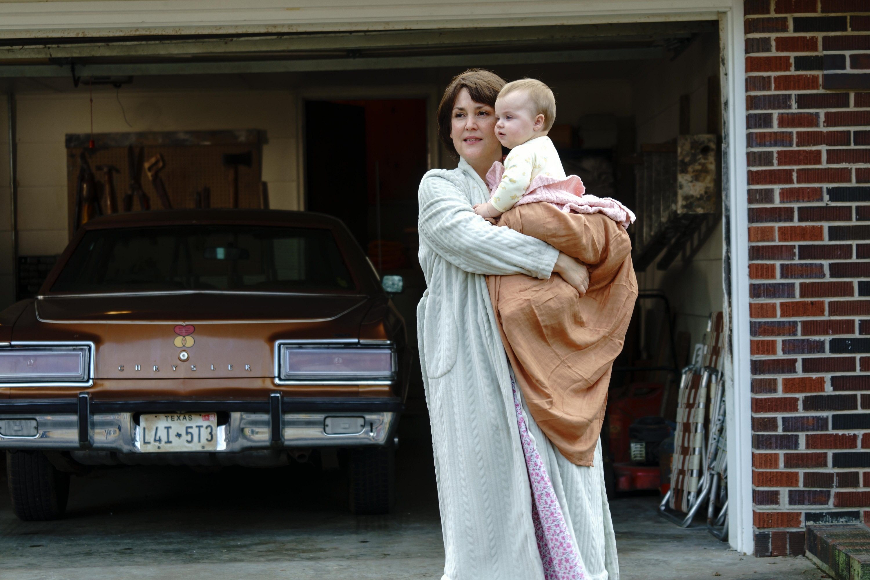 Melanie Lynskey as Betty Gore holding her baby while standing in front of their garage in 'Candy' on Hulu