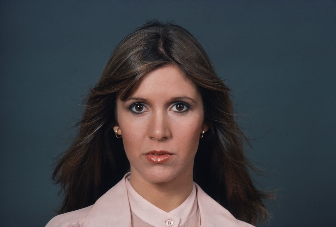 Carrie Fisher wears a pink outfit and stands in front of a gray background.