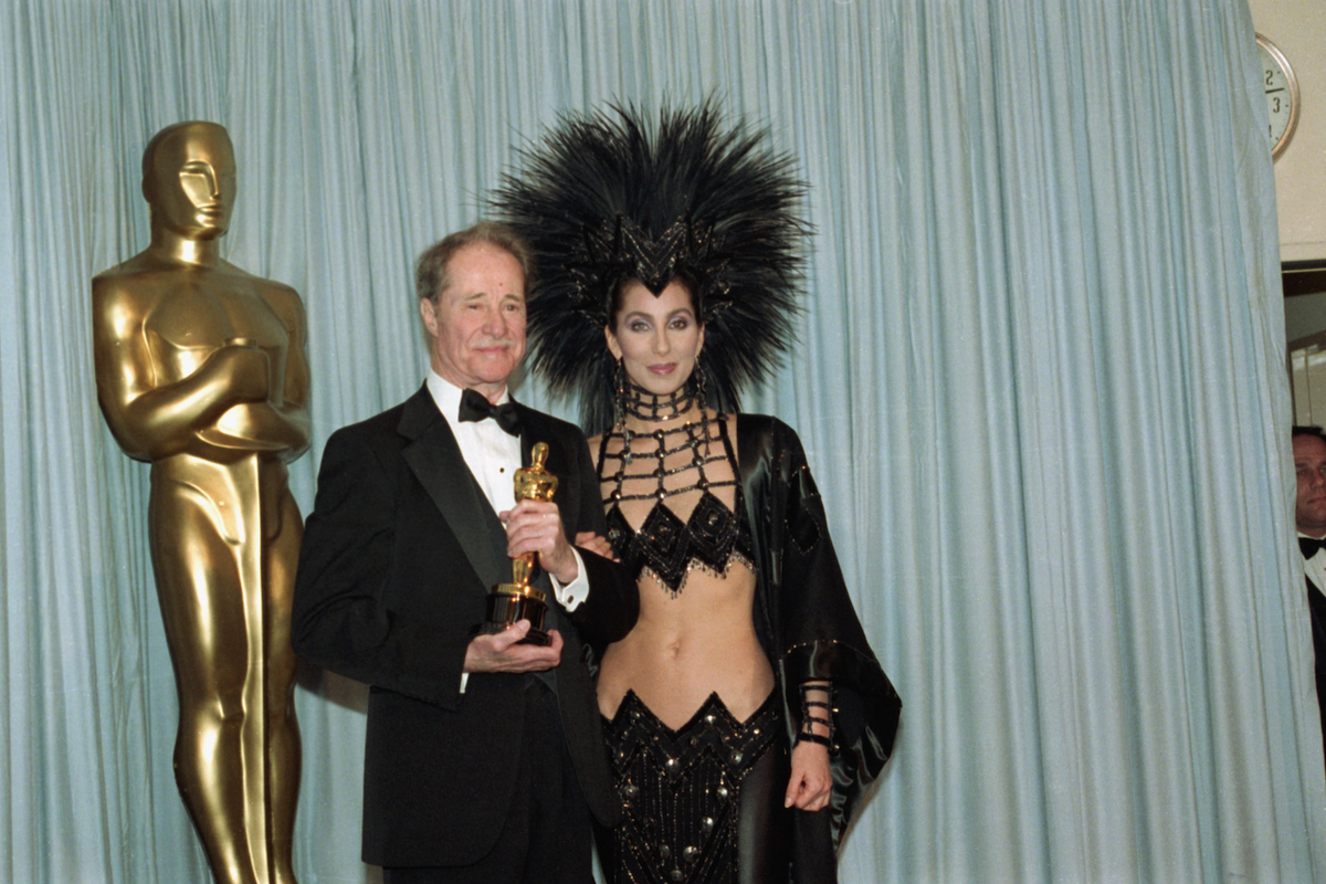 Don Ameche holds the Best Supporting Actor Oscar award for his role in Cocoon, with award presenter Cher, who is wearing an unusual Bob Mackie evening gown with a feather headdress