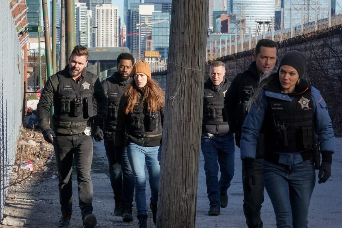 Patrick Flueger, LaRoyce Hawkins, Marina Squerciati, Jason Beghe, Jesse Lee Soffer, and Tracy Spiridakos in Chicago P.D. Season 9. The team walks wearing bullet-proof vests.