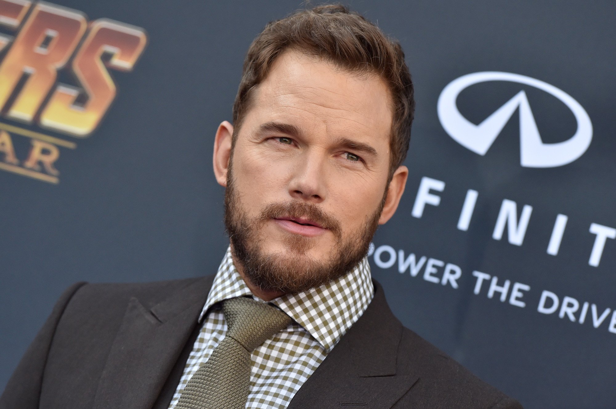 'Guardians of the Galaxy' star Chris Pratt, who appears in 'Thor: Love and Thunder' alongside Chris Hemsworth. The image shows him on the red carpet for 'Avengers: Infinity War.' He's wearing a checkered shirt, suit jacket, and tie.