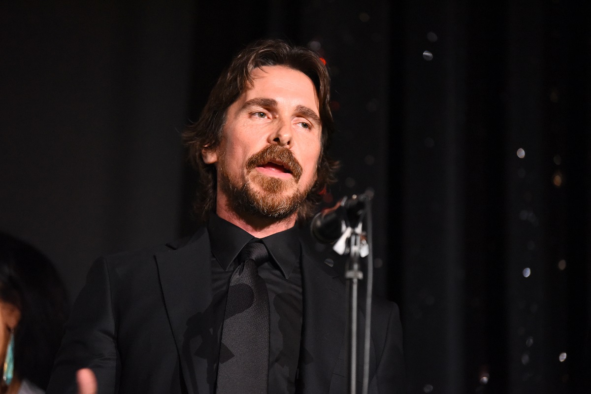 Christian Bale speaking into a microphone while wearing a black suit.