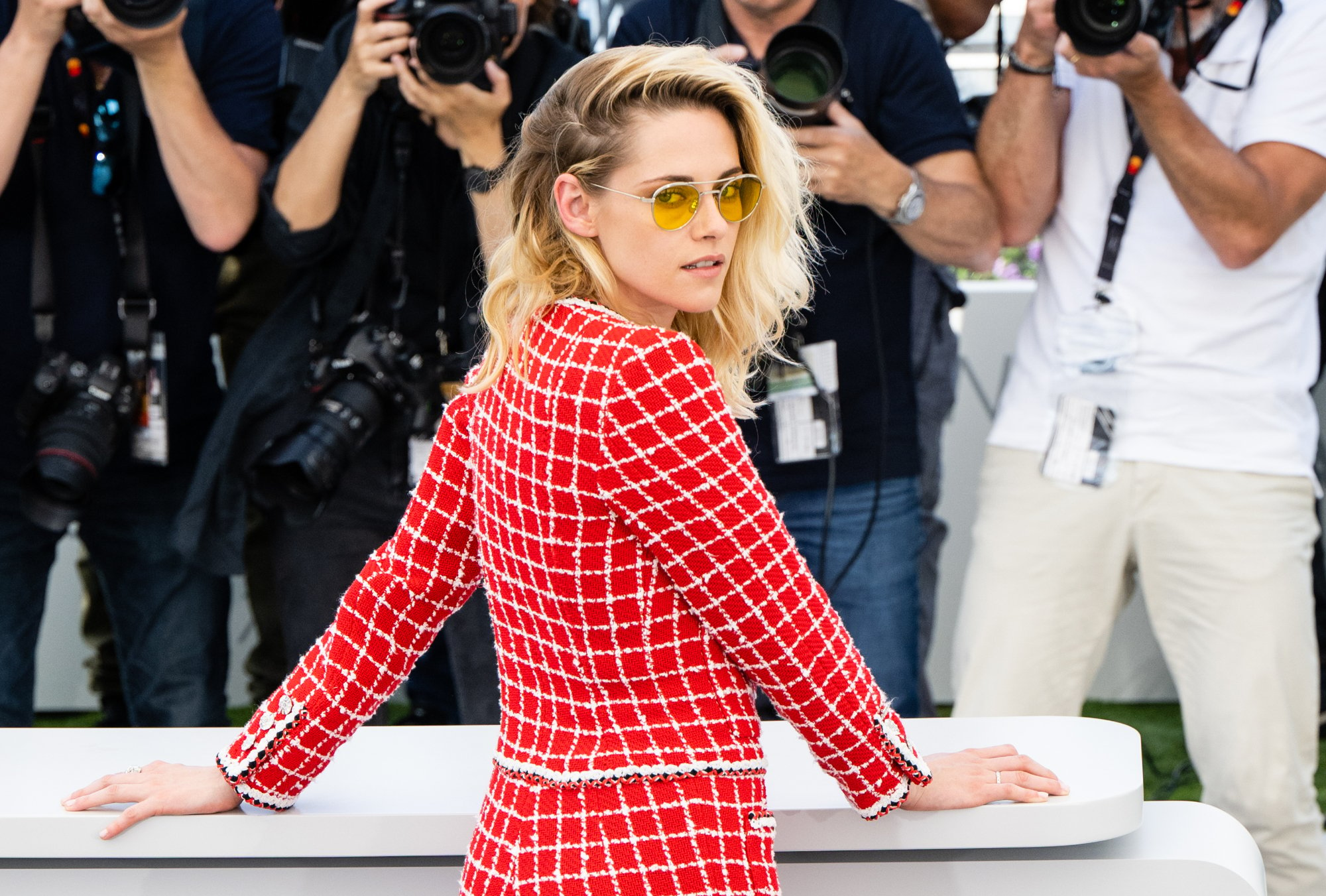 'Crimes of the Future' actor Kristen Stewart wearing a red and white outfit with yellow sunglasses. She has her hands on a white table, looking over her shoulder at the camera.