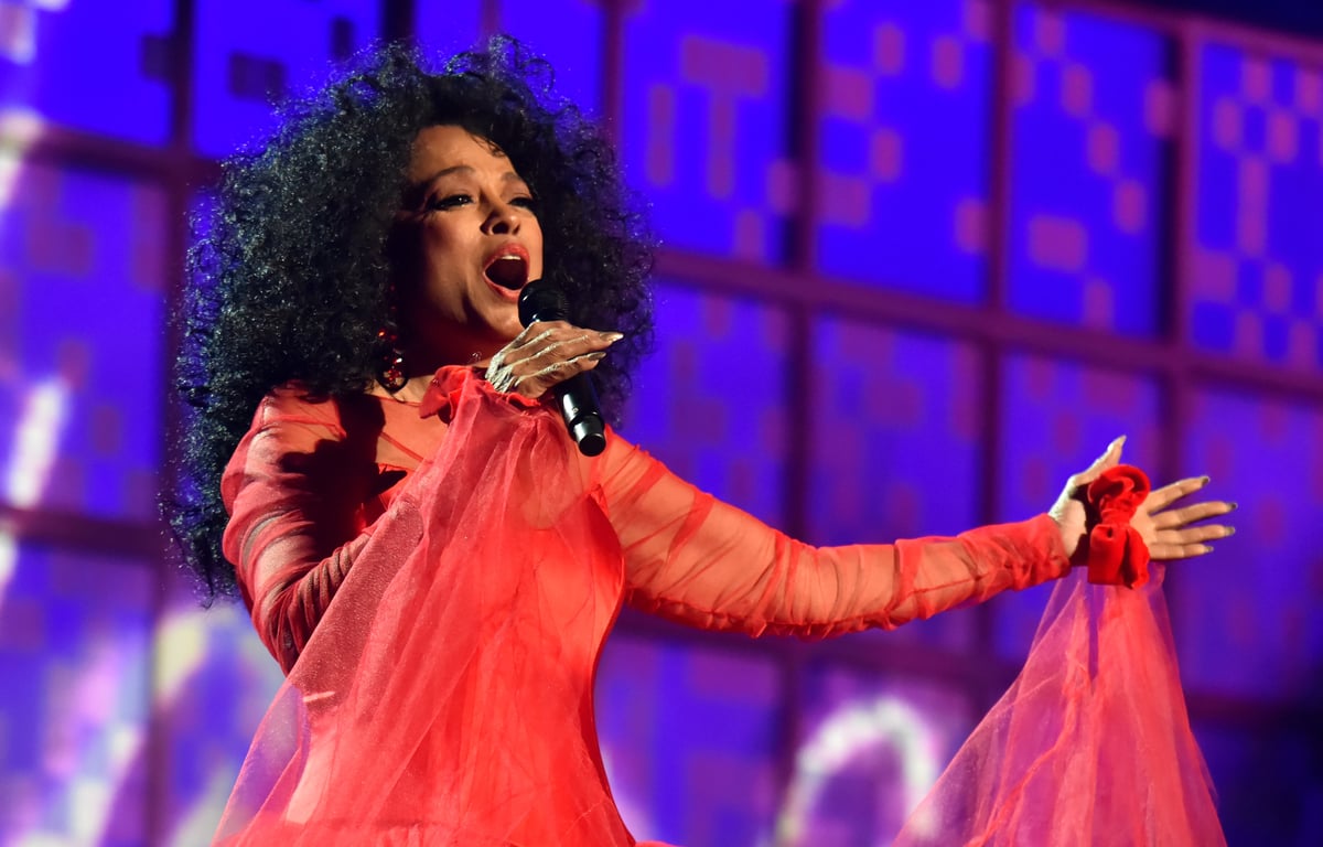 Diana Ross wears a bright red dress while performing in front of a purple backdrop onstage.