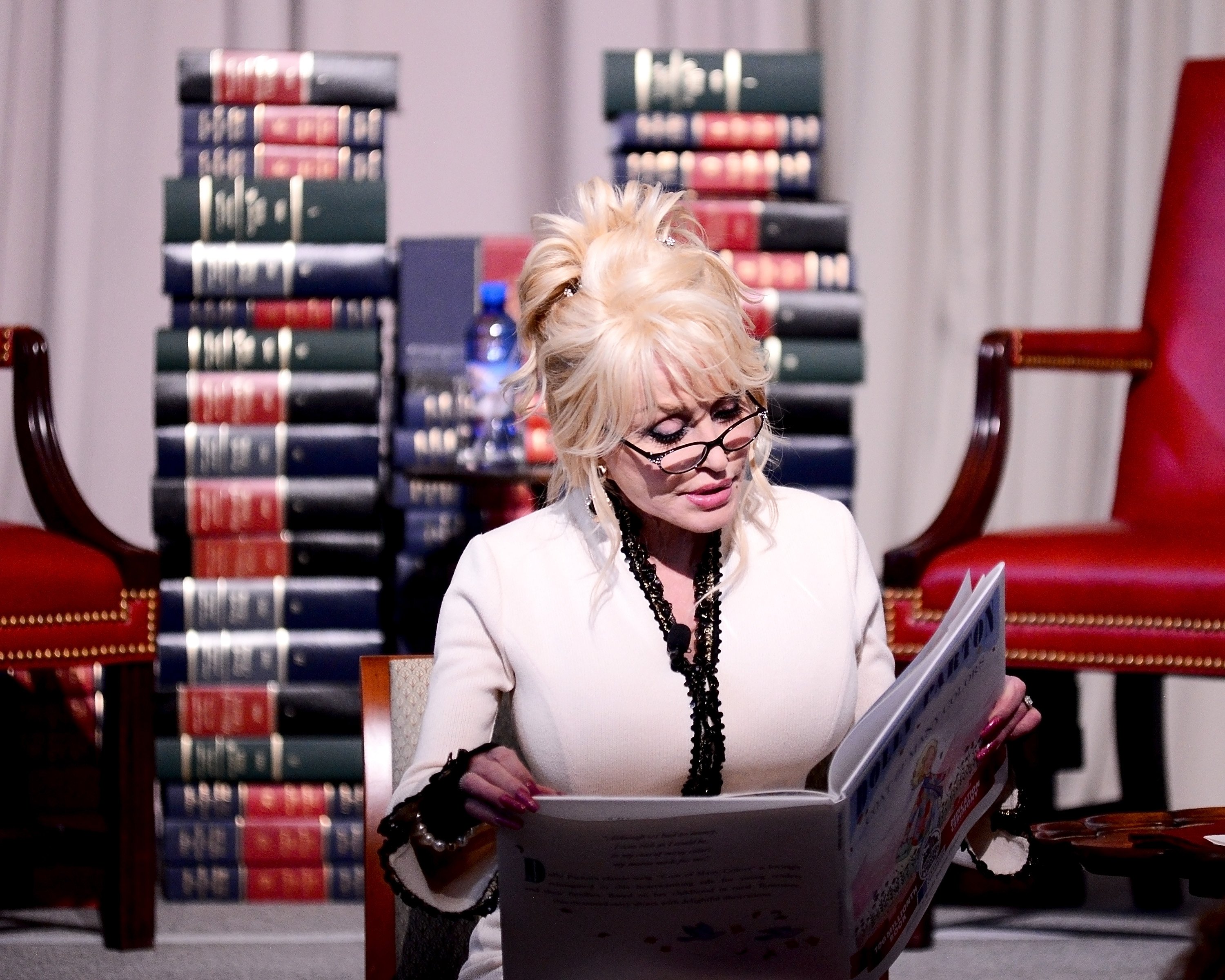 Dolly Parton sits in front of stacks of books and reads from a children's book.