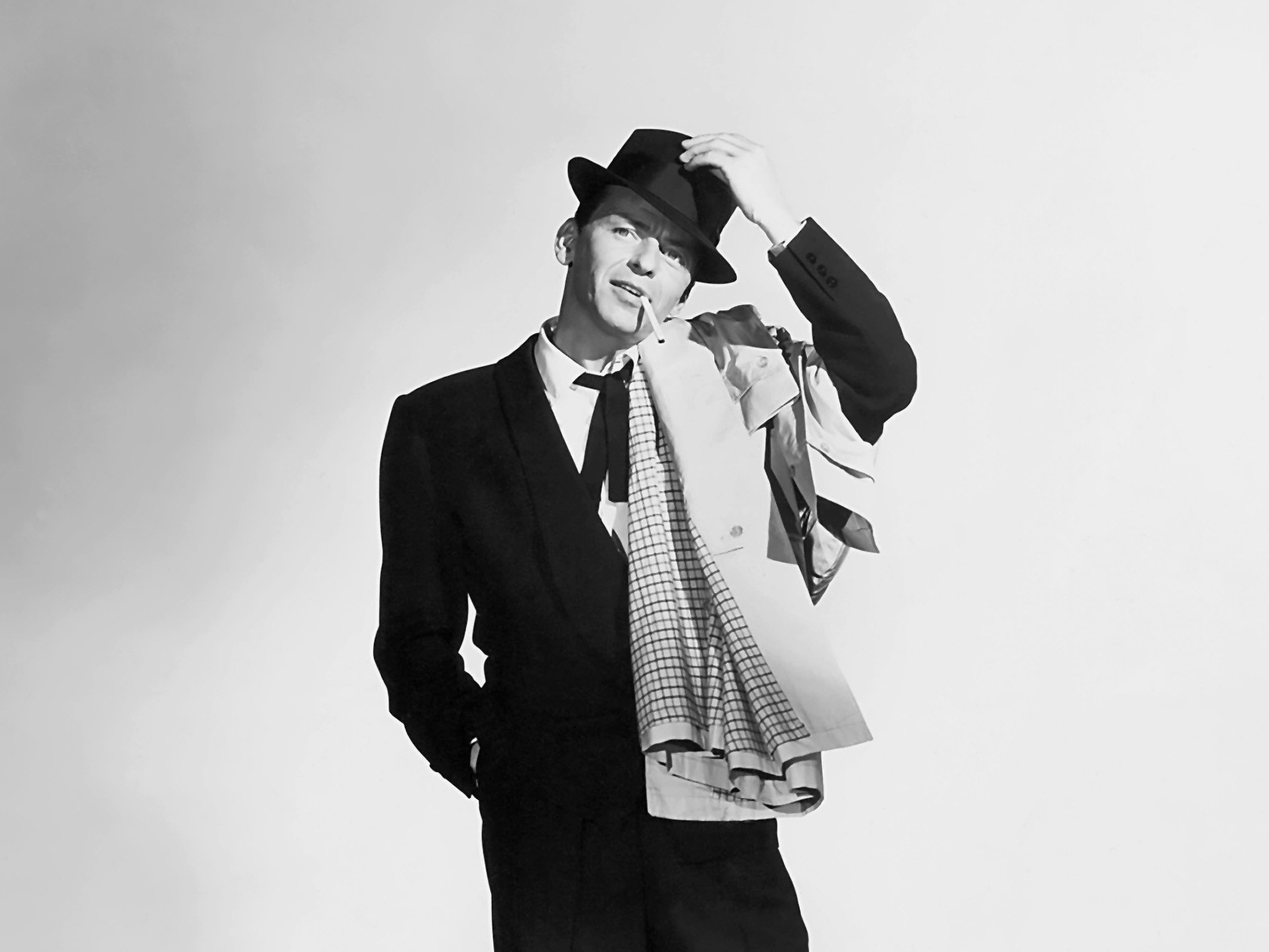 Frank Sinatra wears a suit and hat and hangs his jacket over his shoulder. He has a cigarette in his mouth.