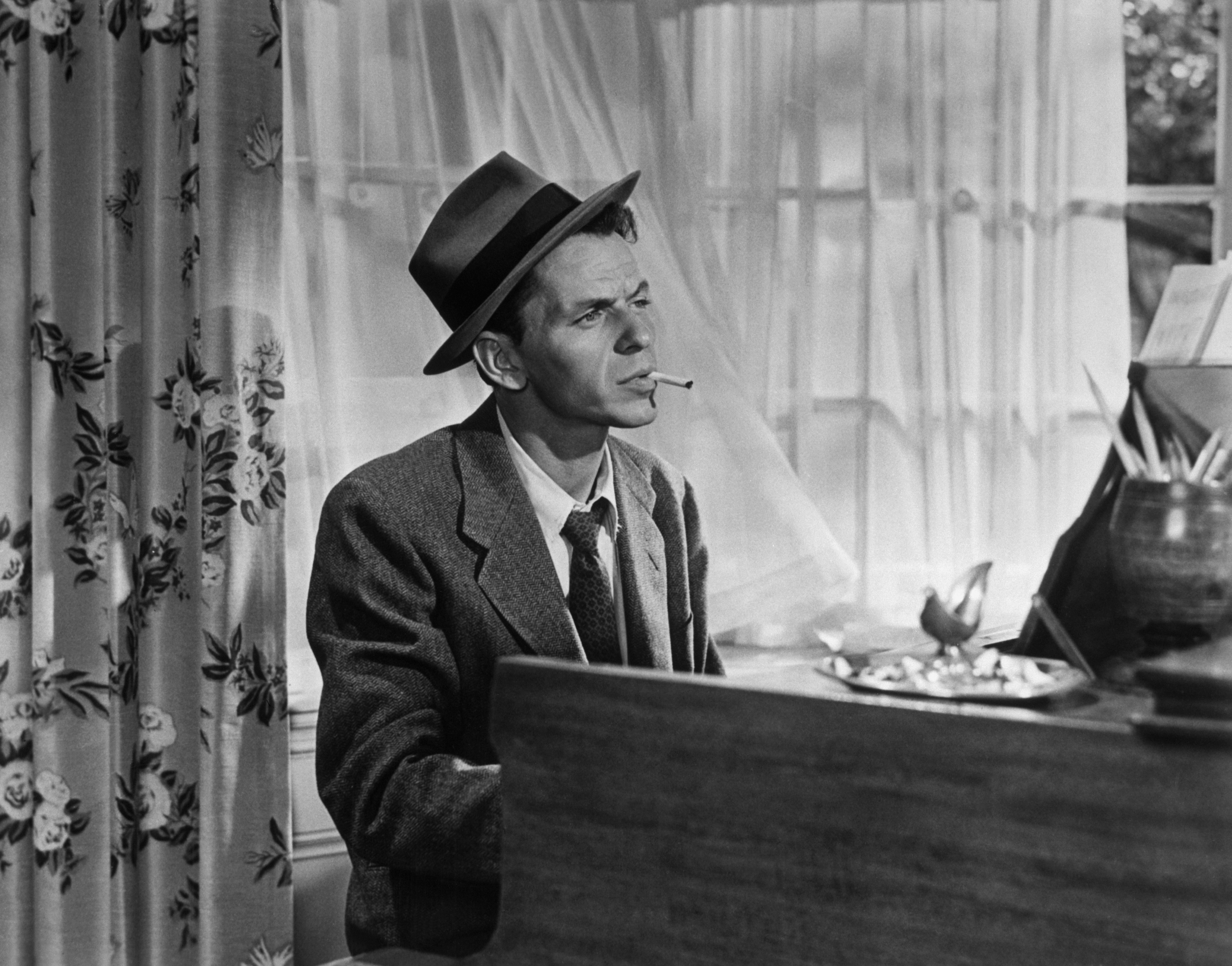 A black and white photo of Frank Sinatra sitting at a piano with a cigarette in his mouth.