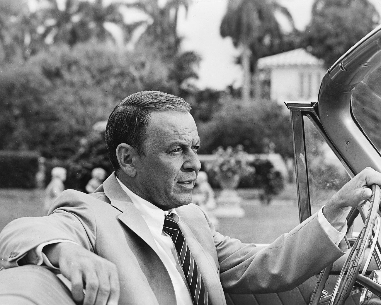 The singer and actor Frank Sinatra wears a suit and sits behind the wheel of a convertible. 