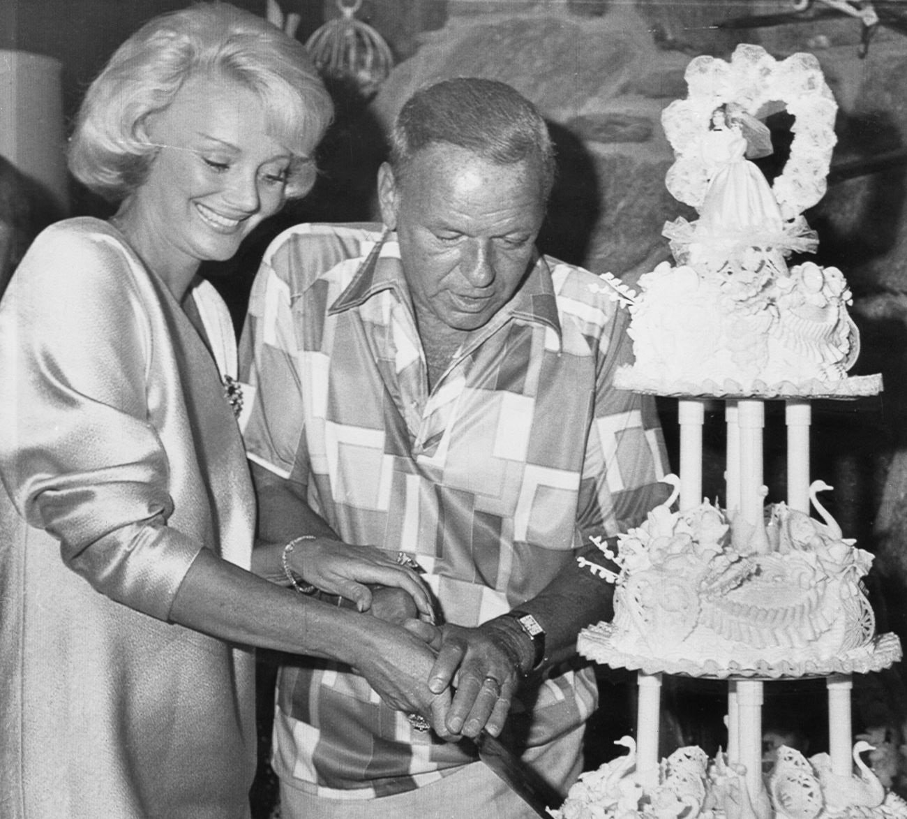 A black and white photo of Frank Sinatra and Barbara Sinatra cutting into their wedding cake together.
