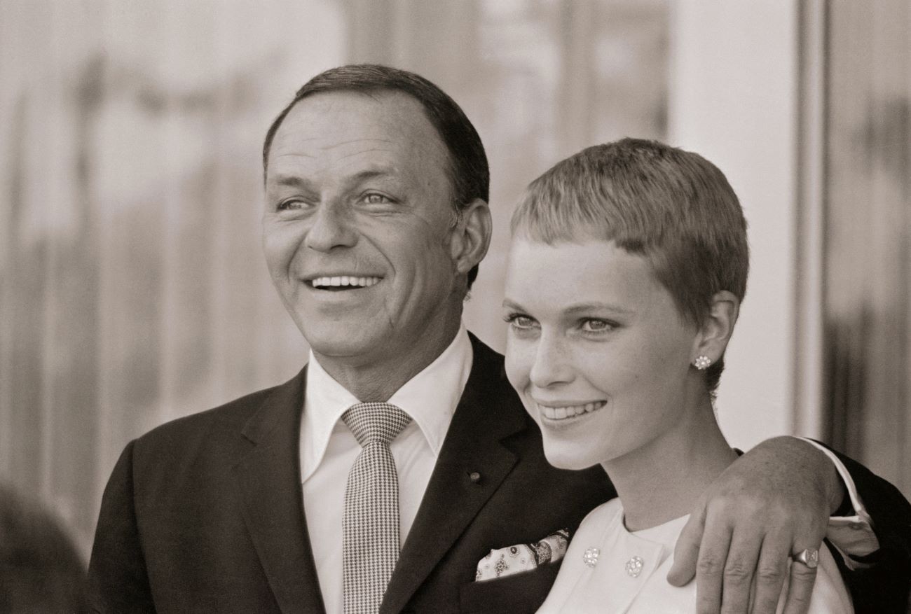 A black and white photo of Frank Sinatra standing with his hand around Mia Farrow's shoulders.