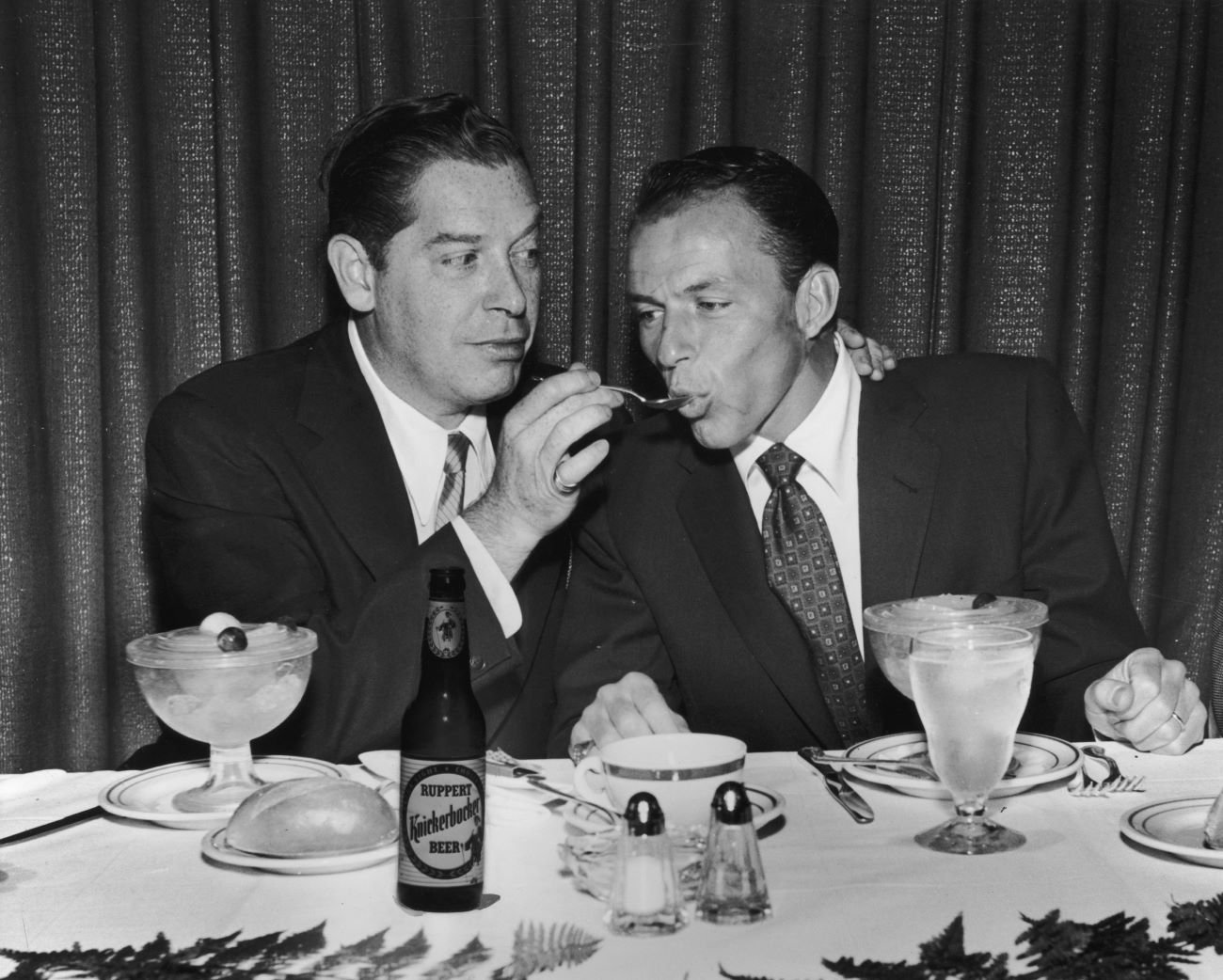 Comedian Milton Berle feeds Frank Sinatra a spoonful of fruit at a restaurant.