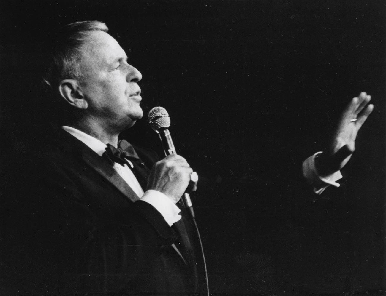 A black and white photo of singer Frank Sinatra wearing a tuxedo and holding a microphone.
