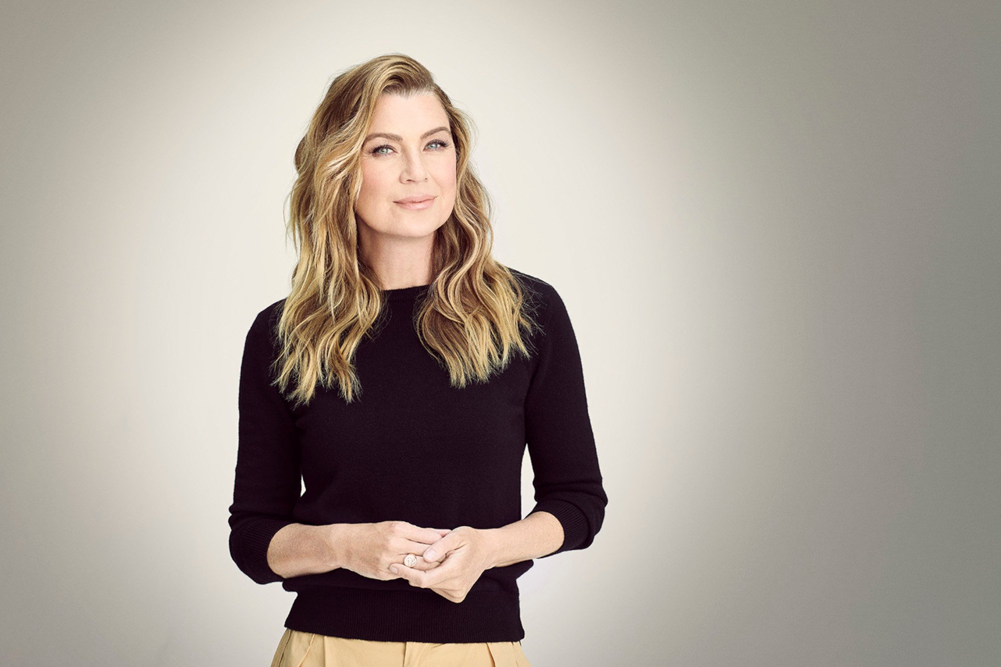 'Grey's Anatomy' star Ellen Pompeo. She's wearing a black shirt, beige pants, and standing in front of a white backdrop. She's smiling at the camera, and her blonde hair is down.