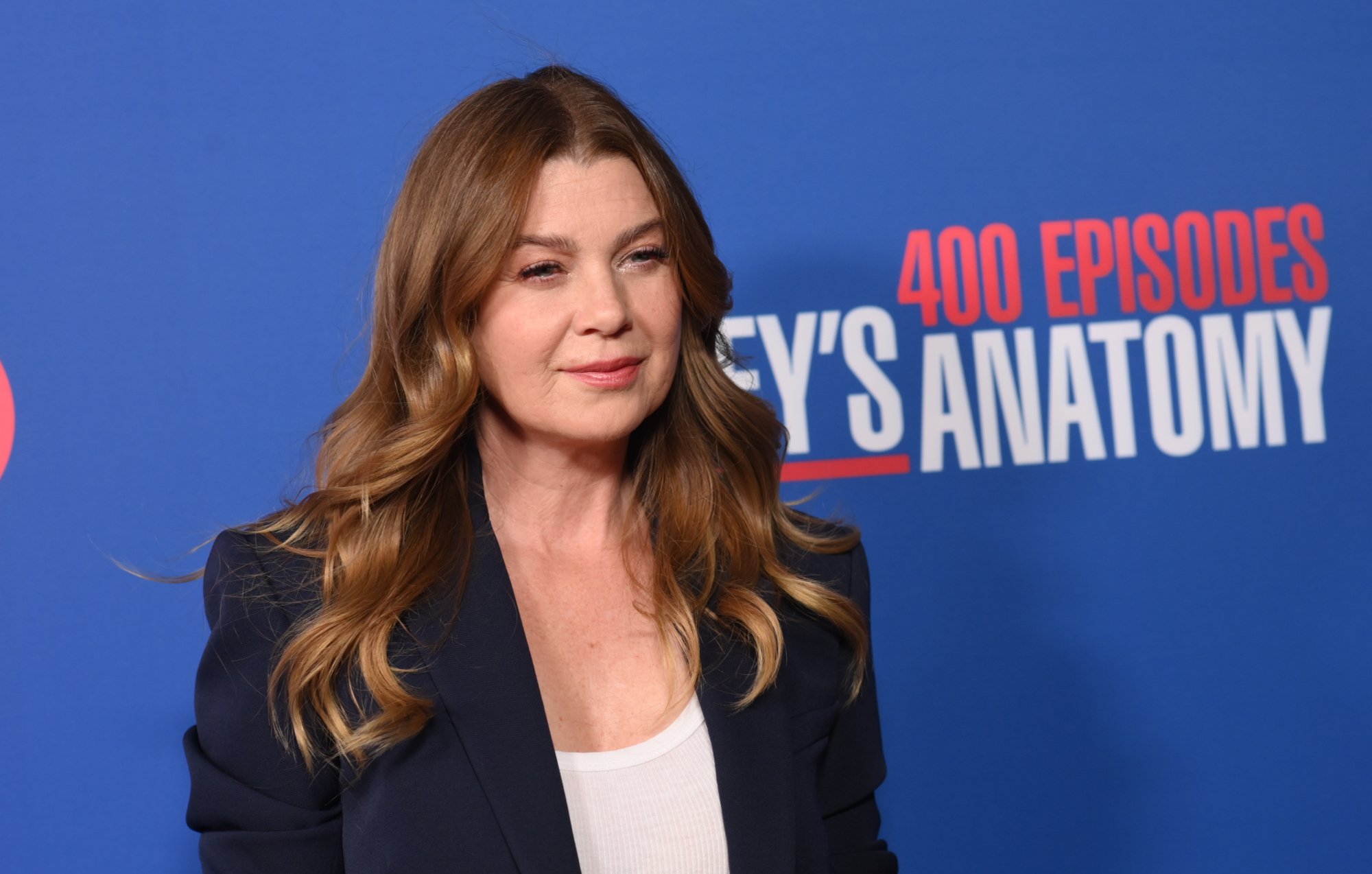 'Grey's Anatomy' star Ellen Pompeo standing in front of a wall commemorating the show's 400th episode. She's wearing a white shirt and blue blazer, and her hair is down.