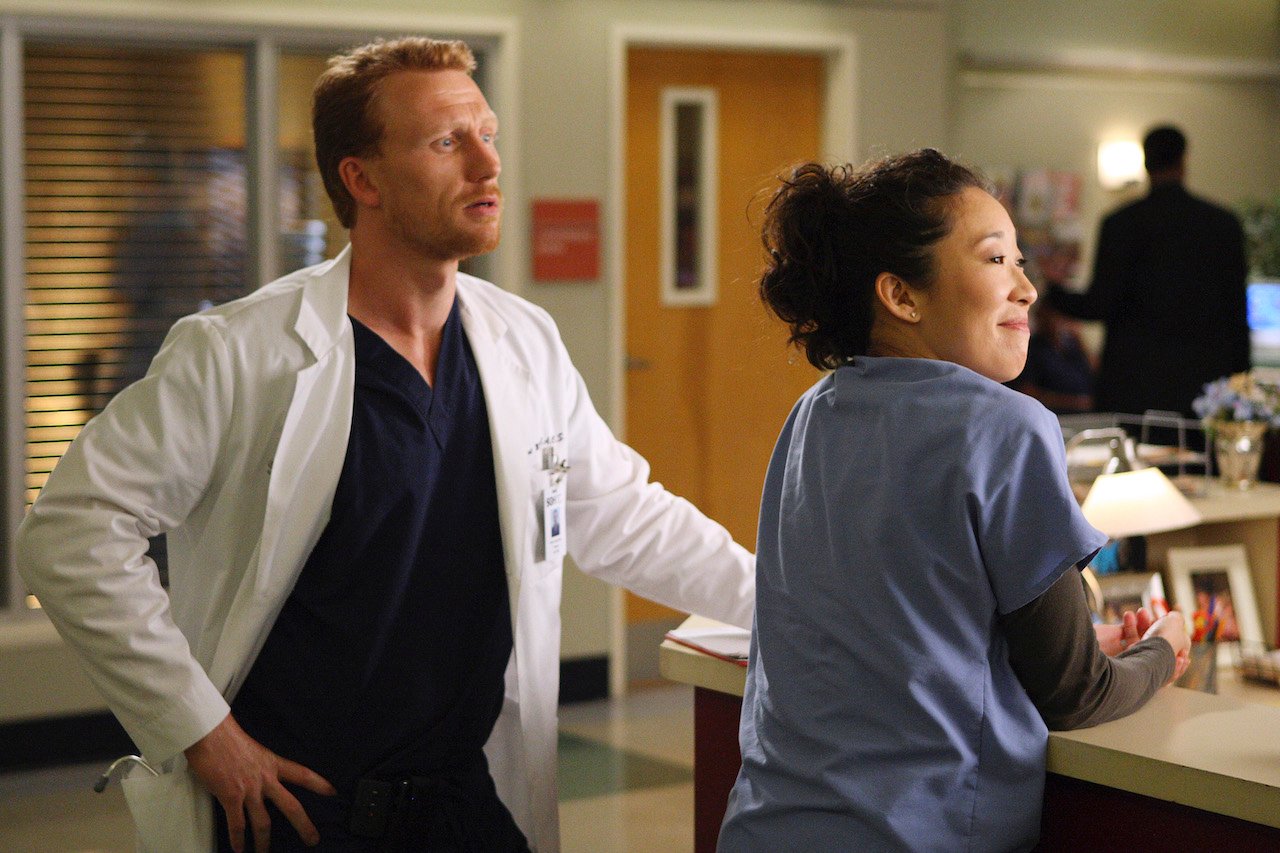 Kevin McKidd as Owen Hunt and Cristina Yang as Sandra Oh stand next to each other in the hospital on 'Grey's Anatomy'.