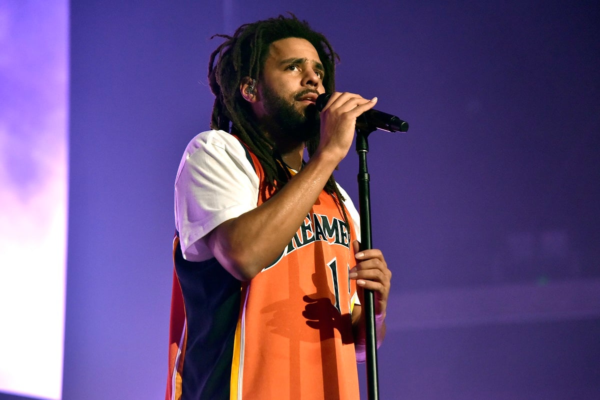 J. Cole speaking into a microphone while wearing an orange jersey.