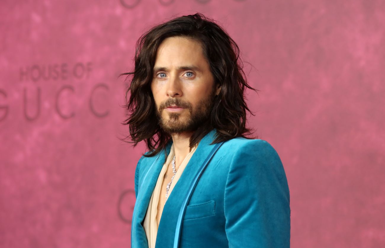 Jared Leto wears a blue suit against a pink background.