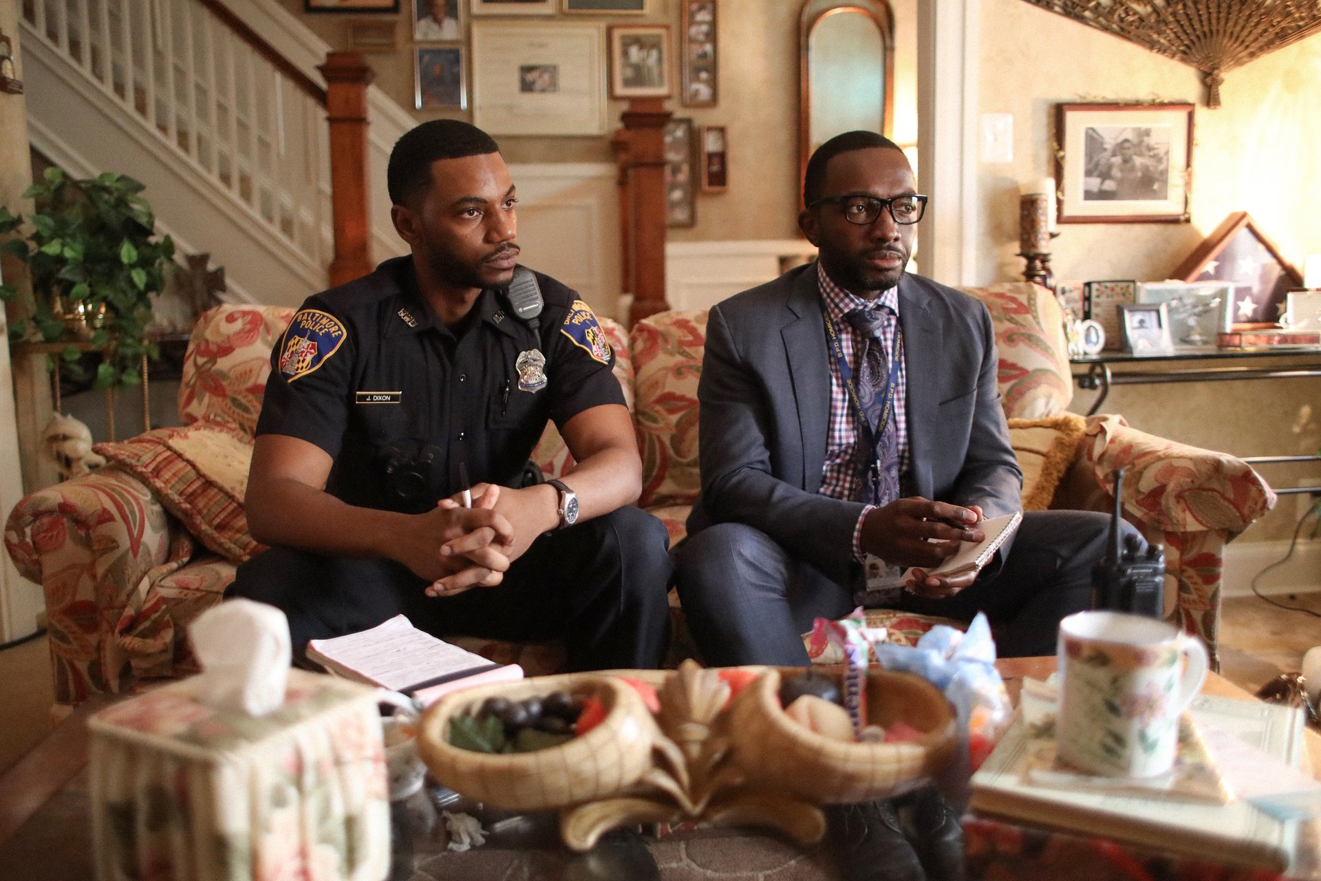 'The Wire' actor Jermaine Crawford as Jaquan Dixon wearing a cop uniform and Jamie Hector as Sean Suitor wearing a suit sitting on a couch in 'We Own This City'