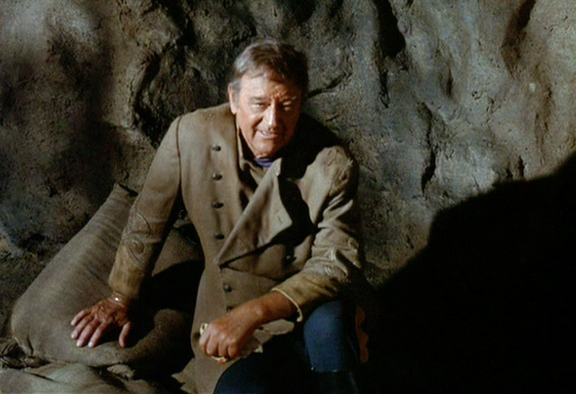 John Wayne as Colonel Cord McNally in 'Rio Lobo,' who isn't afraid of a fight. He's wearing his Western costume and resting back against a rock wall with his arm resting on his knee.