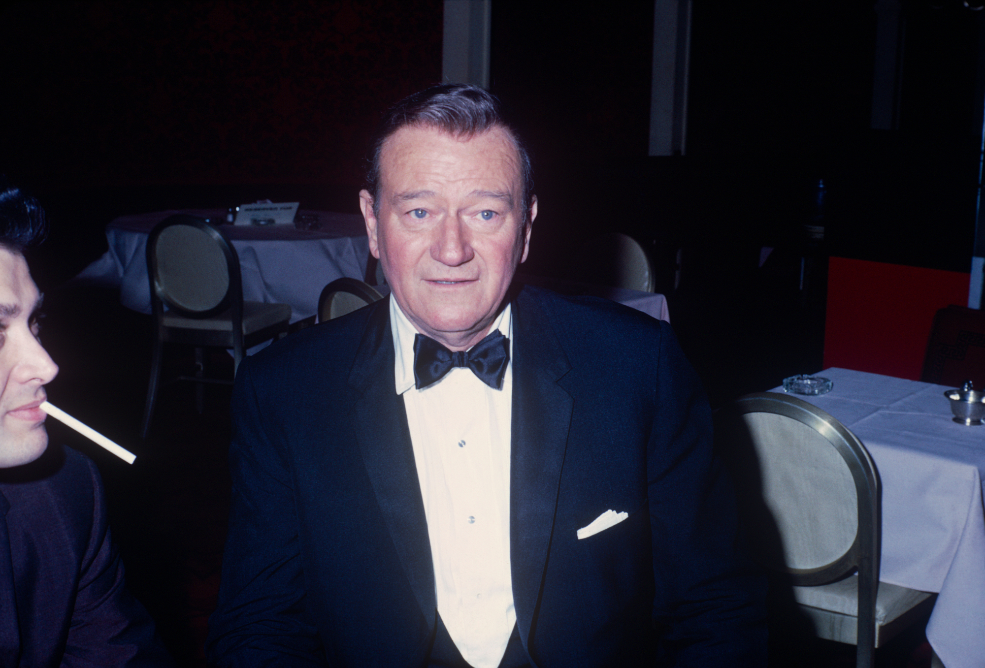 John Wayne, who had negative views on the celebration of gay rights, wearing a black-and-white tuxedo with white table-clothed tables in the background