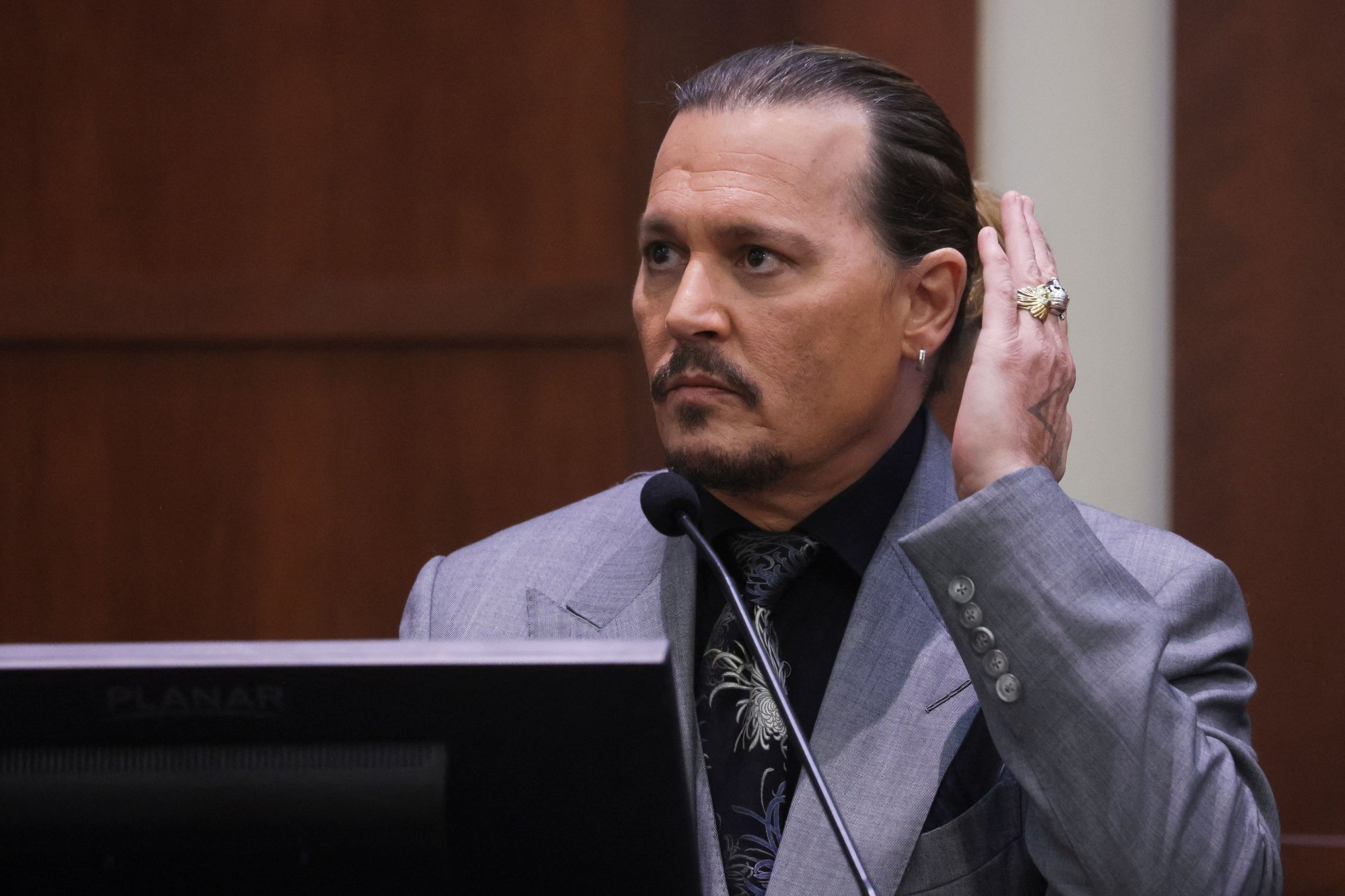 Johnny Depp testifying about alleged drunk moment wearing a suit in front of a microphone with his hand up by his ear