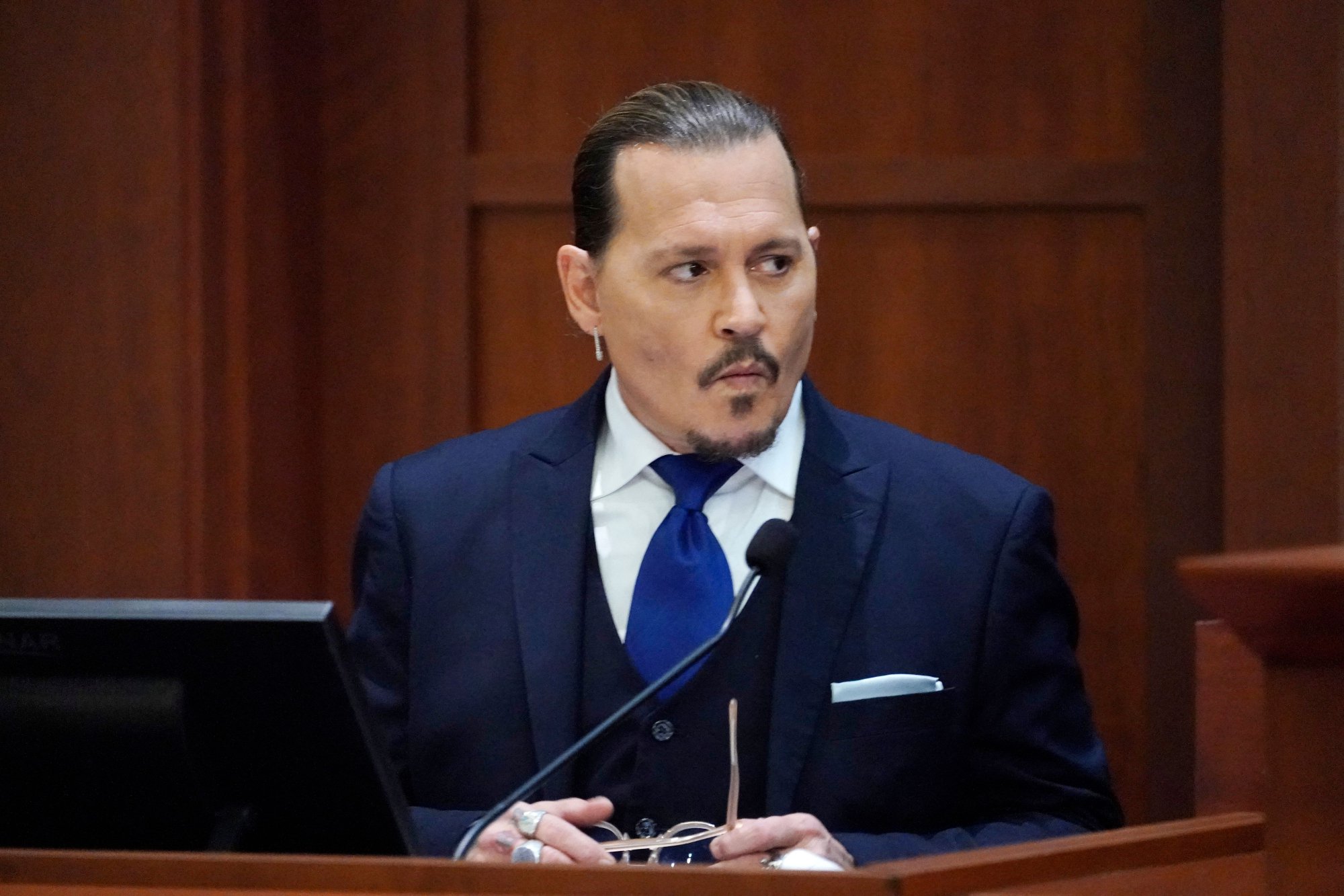 Johnny Depp testifies, wearing a blue suit and holding his sunglasses, at his trial versus Amber Heard. Depp joked about hearsay while on the stand.