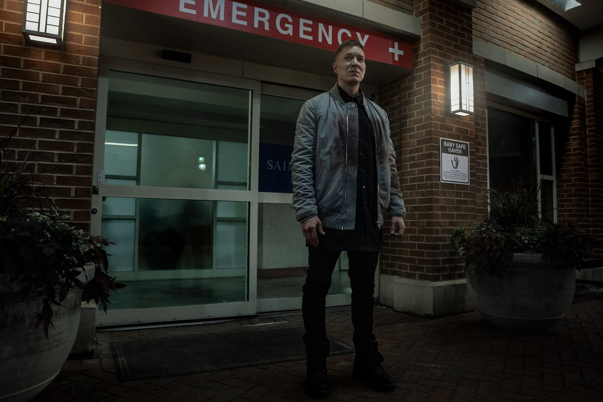 Joseph Sikora appears in a scene from 'Power Book IV: Force' as his character Tommy Egan. He's wearing a green jacket over a black shirt and jeans with matching shoes while standing outside of an emergency room.
