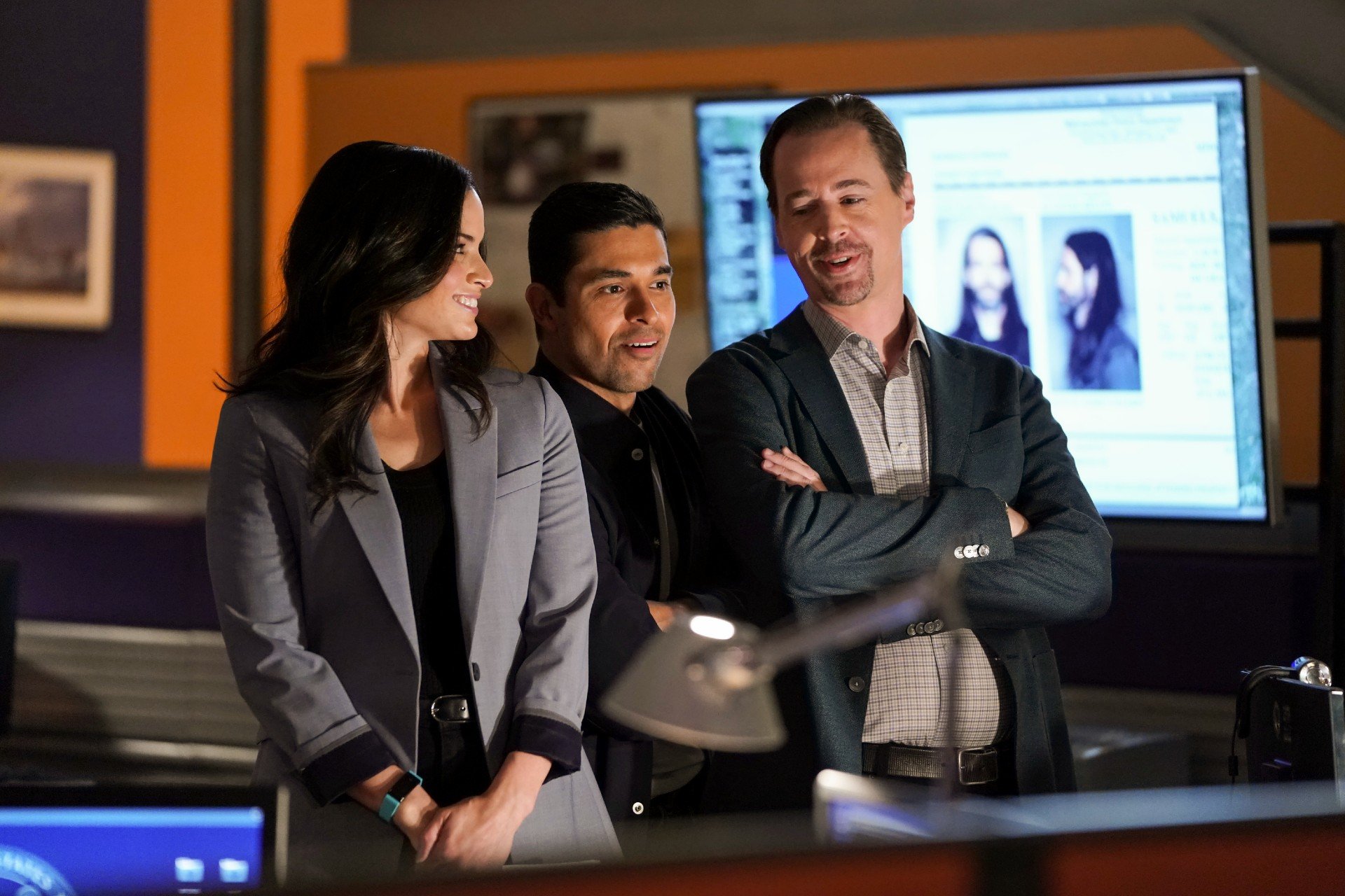 The NCIS team stands together in the office and smiles.