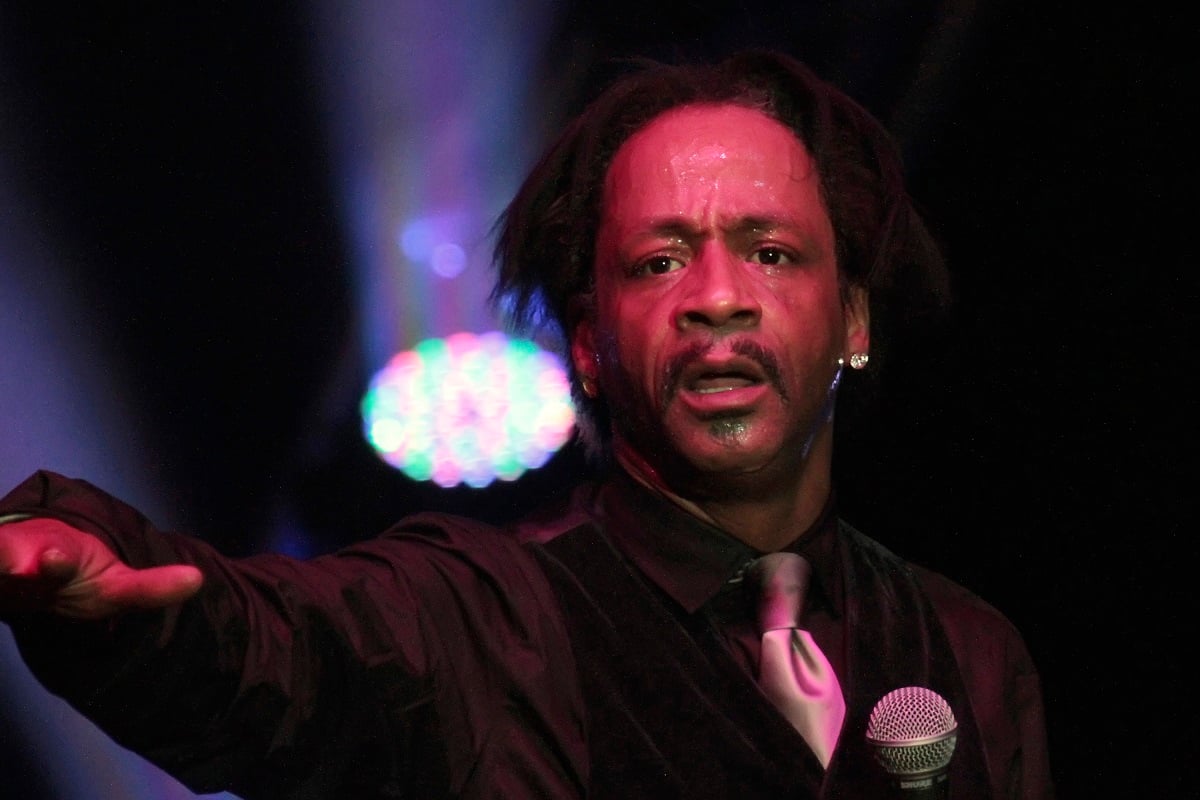 Katt Williams holding a microphone while wearing a black suit.