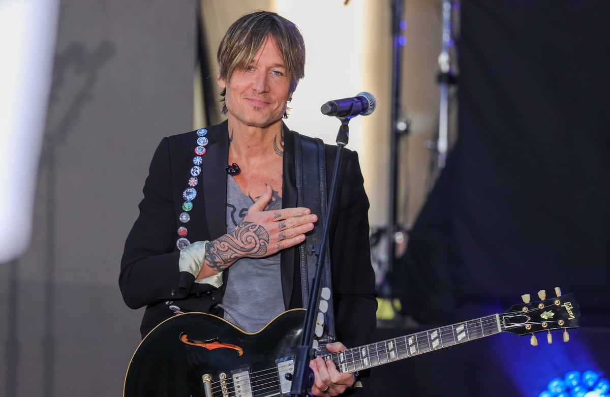 Keith Urban holds his guitar in one hand and has his other hand on his heart as he smiles on stage.