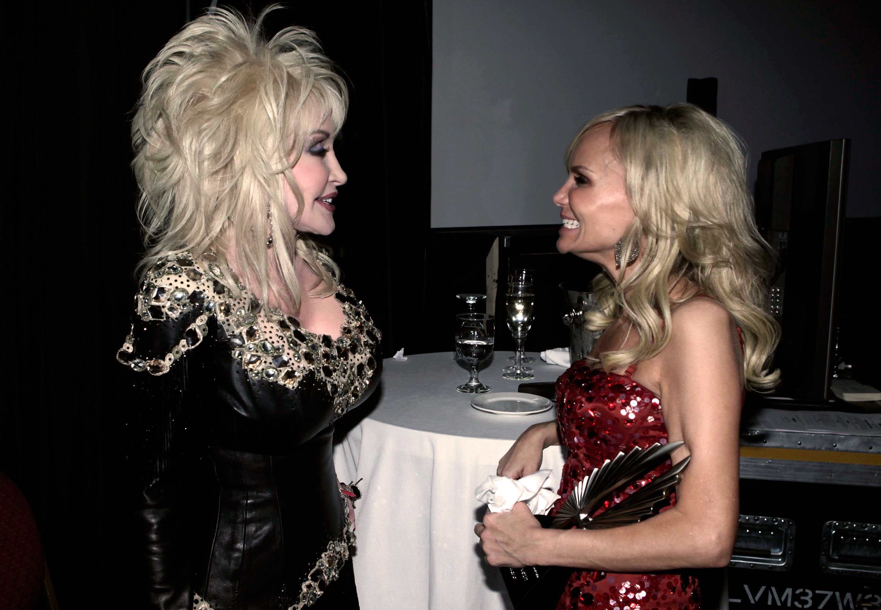 Dolly Parton and Kristin Chenoweth look at each other in front of a dinner table.