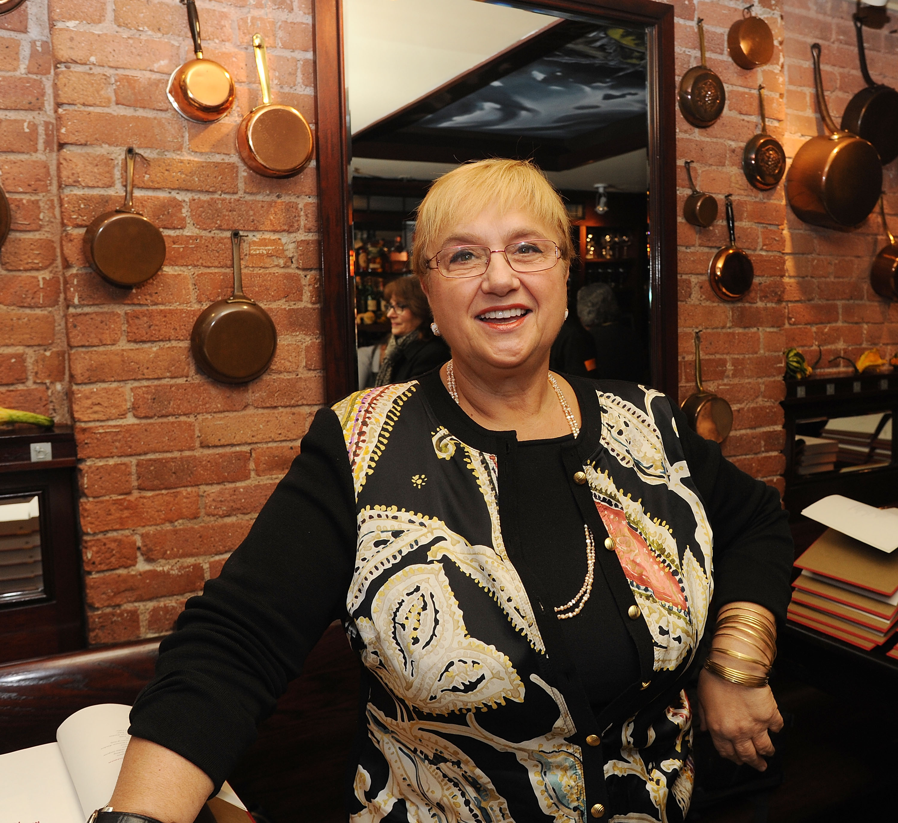 Lidia Bastianich wears glasses and a white blouse as she smiles in this photograph.