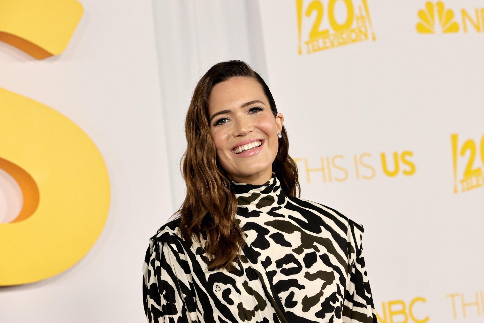 'This Is Us' star Mandy Moore, who plays Rebecca Pearson on the show. She's wearing a white dress with black leopard-spot-like patterns on it. She's smiling and standing in front of a wall with 'This Is Us' written on it.