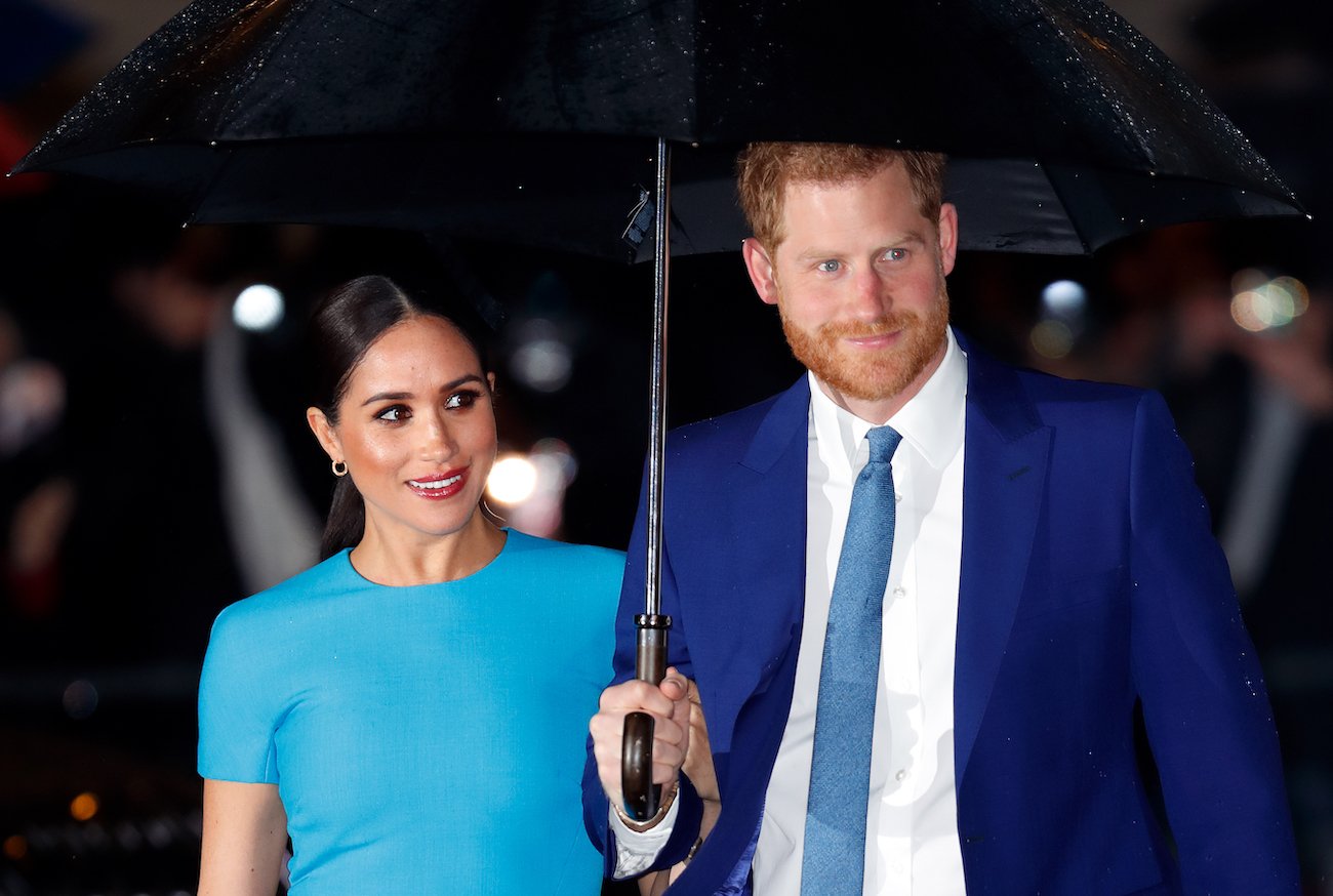 Meghan Markle wearing a light blue outfit and Prince Harry wearing a dark blue suit. Both standing under an umbrella