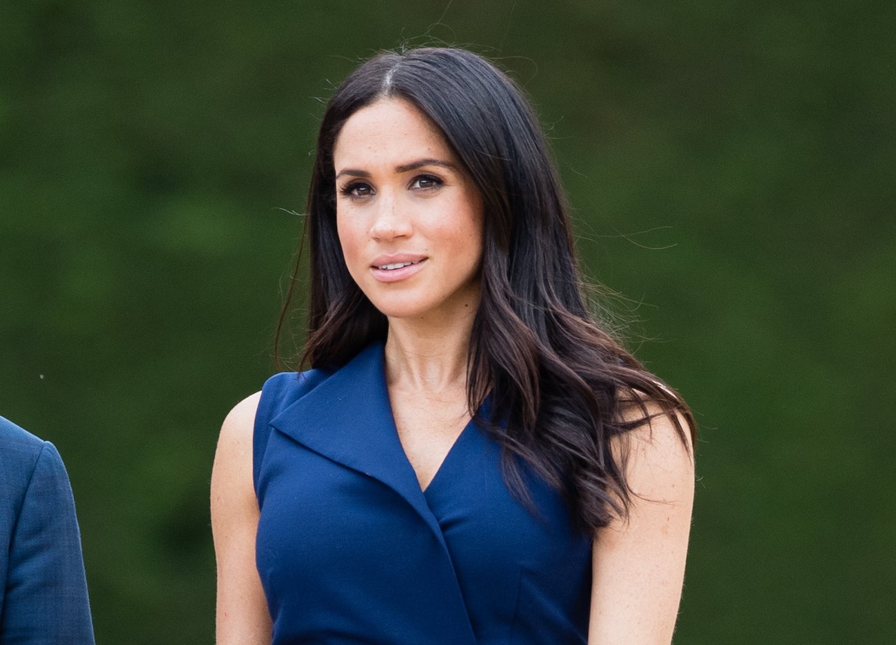 Meghan Markle wearing a sleeveless dark blue dress and looking on