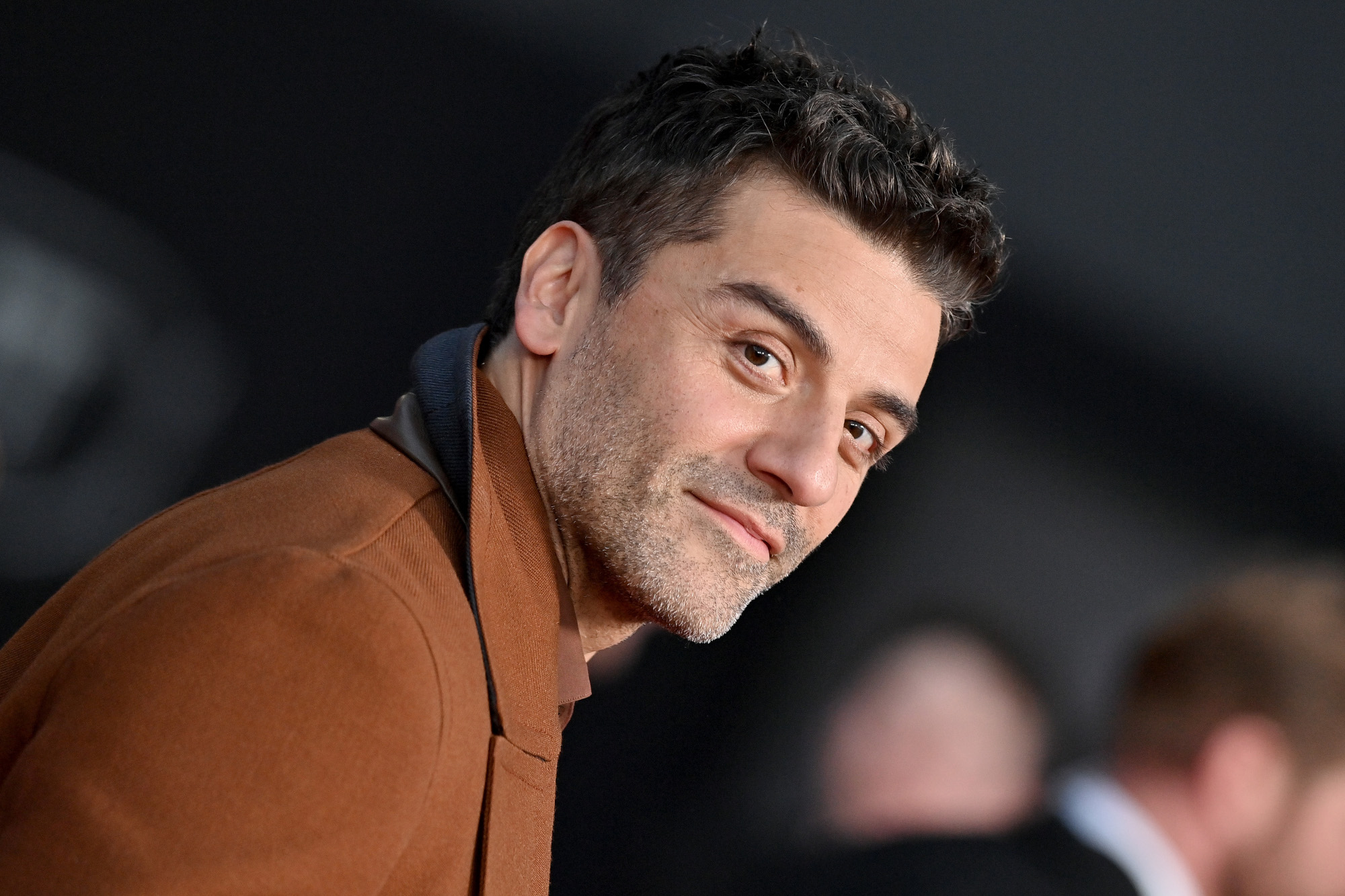 Marc Spector, Steven Grant, and Jake Lockley actor Oscar Isaac at the premiere for Marvel's 'Moon Knight.' He's wearing a brown jacket and smiling at the camera.