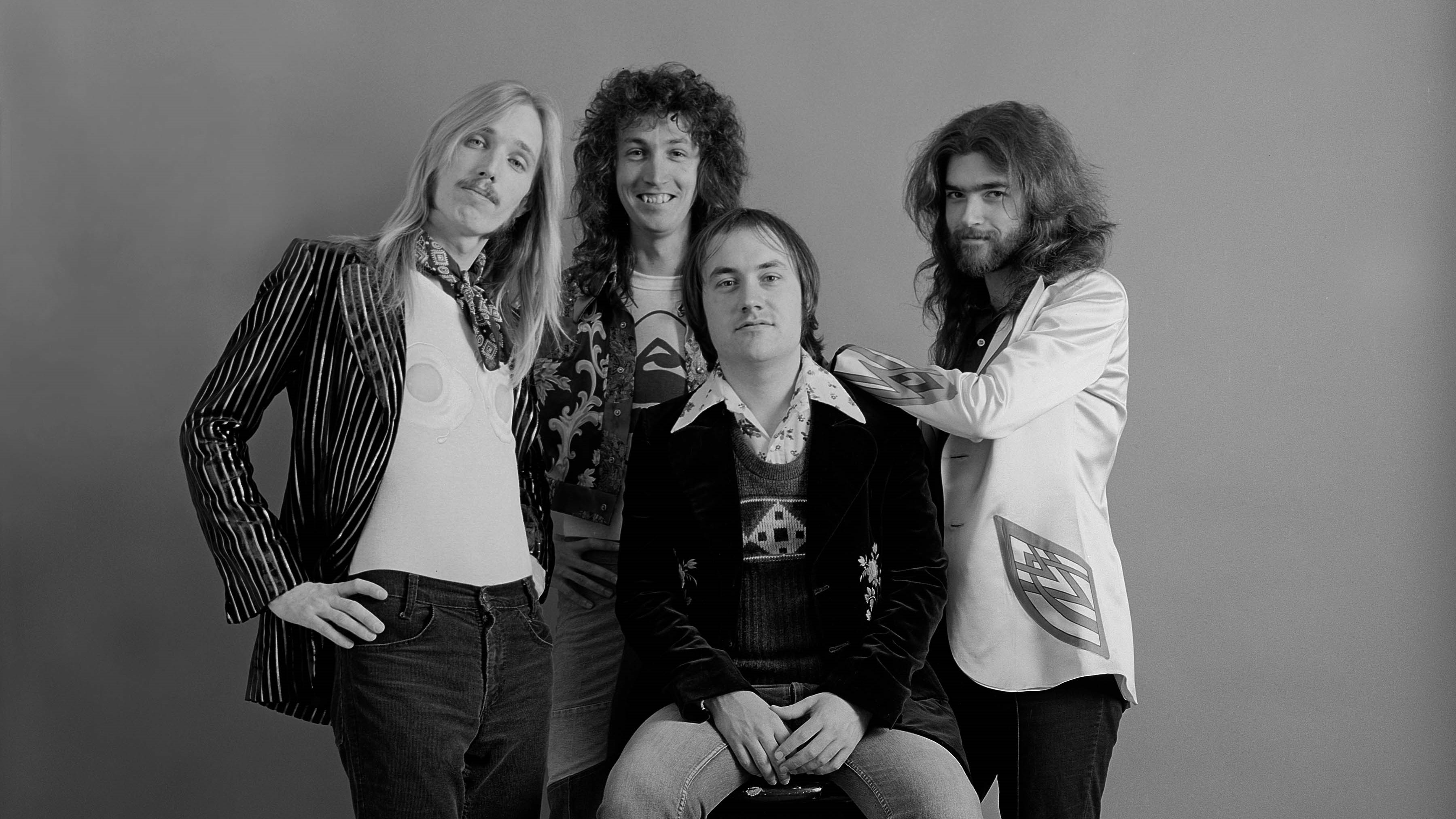 A black and white picture of Tom Petty, Mike Campbell, Randall Marsh, and Tom Leadon of the band Mudcrutch. Marsh sits on a stool and the other men stand around him.