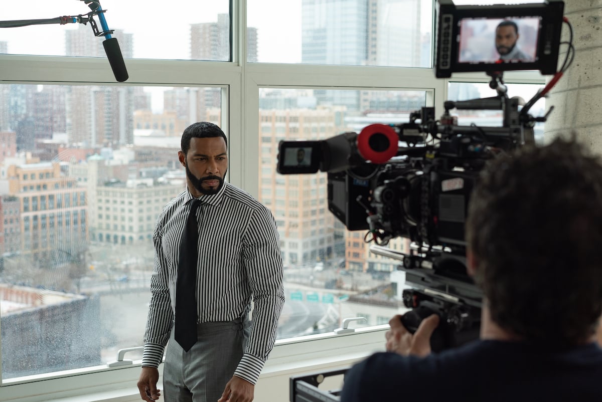 Omari Hardwick films a scene from 'Power' wearing a buttoned up shirt with stripes, a tie, and gray dress pants