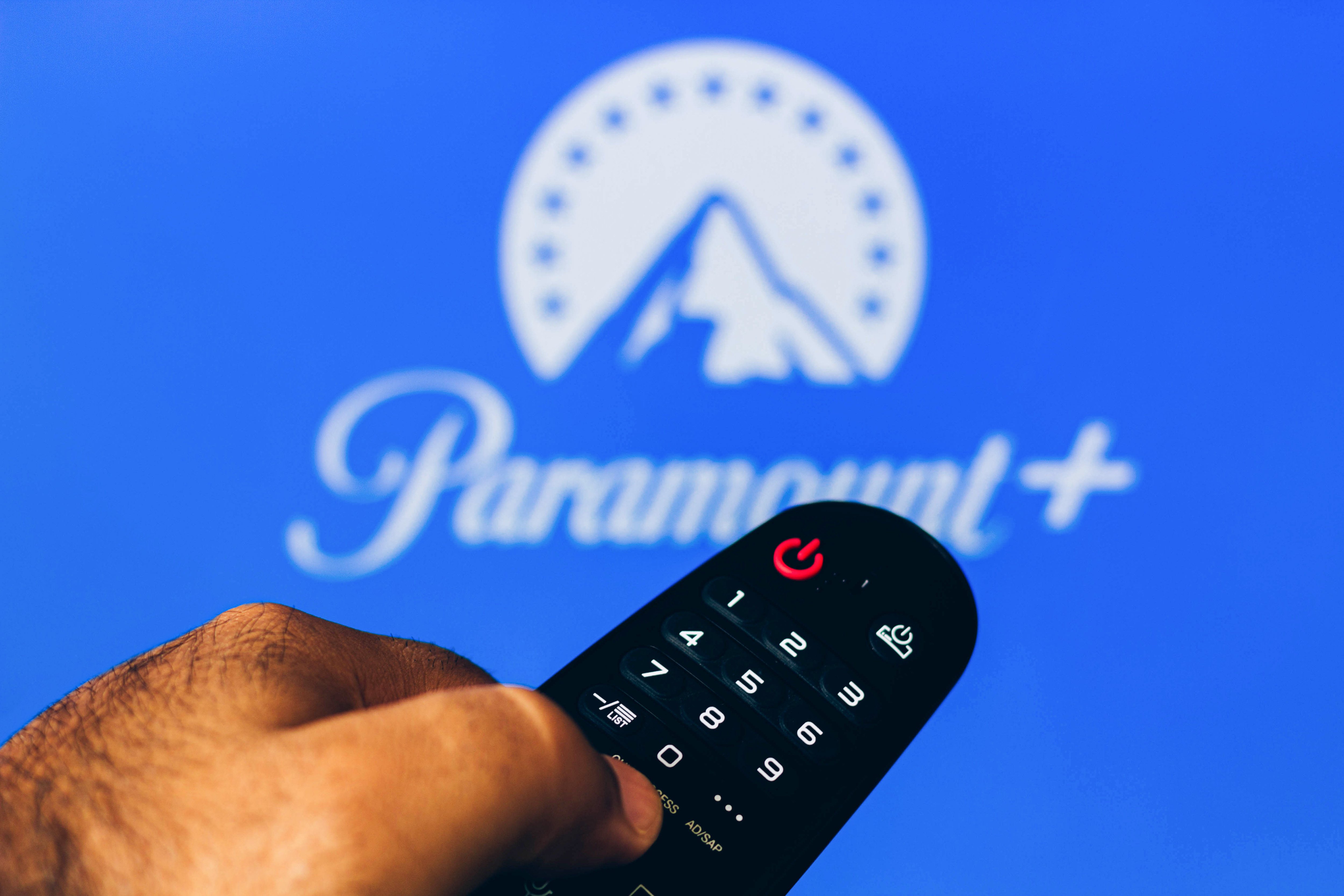 In this photo illustration, a hand holding a TV remote control points to a screen that displays the Paramount Plus logo