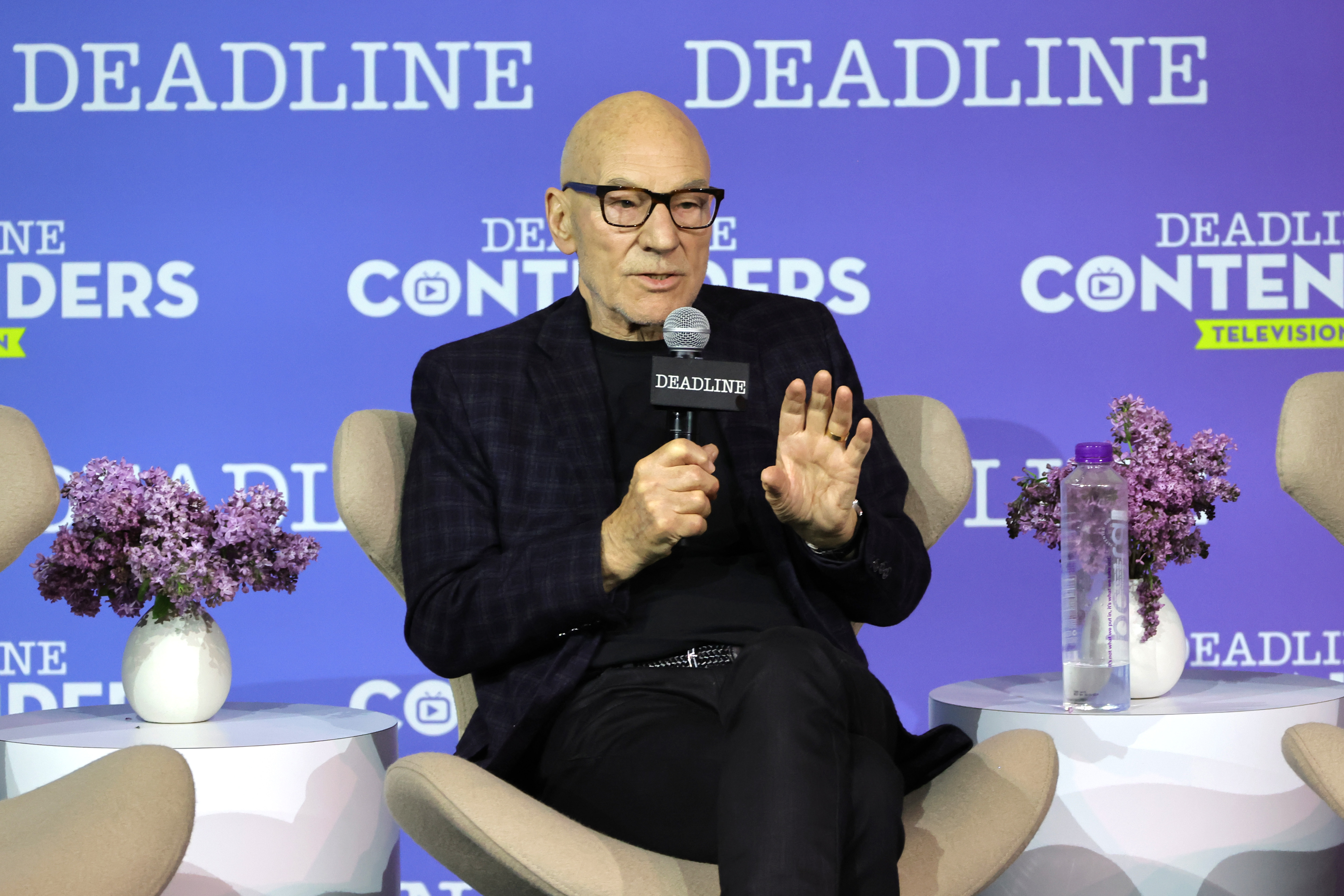 Patrick Stewart, who reprises his role as Professor X in 'Doctor Strange 2,' wears a dark plaid suit over a black shirt and black pants while speaking onstage.