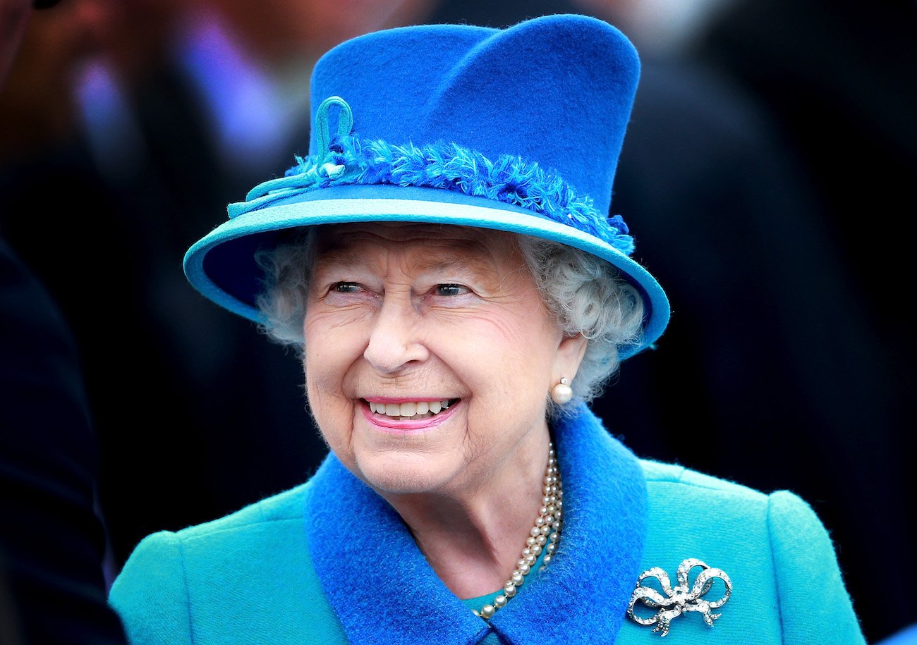 Queen Elizabeth smiling while wearing a blue outfit and a blue hat