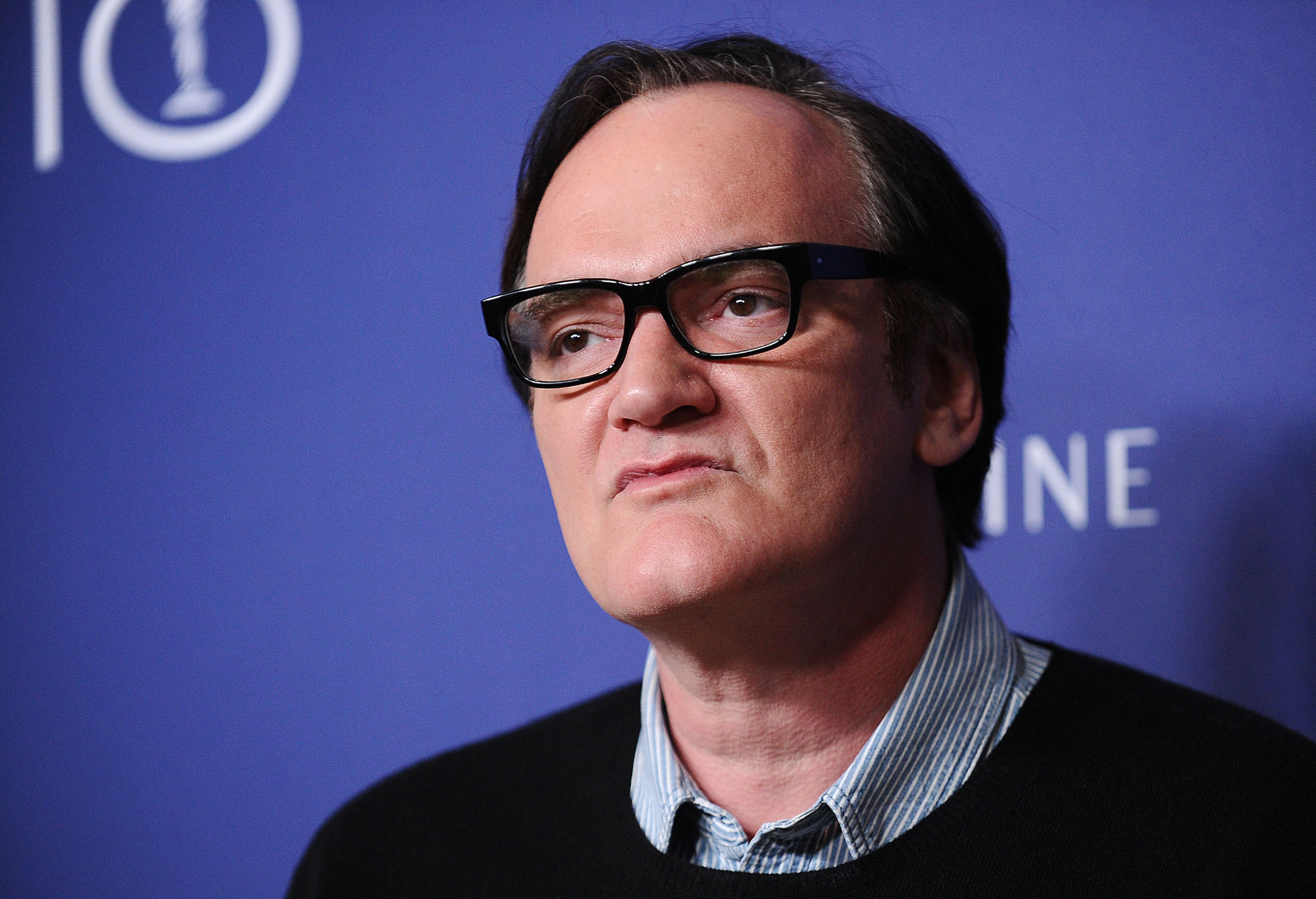 Quentin Tarantino, who collaborates with recurring actors, wearing glasses and a collared shirt in front of a blue step and repeat