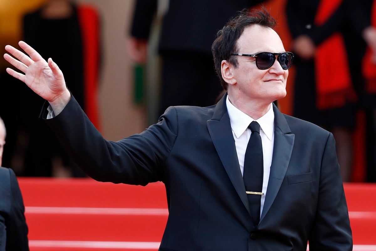 Quentin Tarantino posing while wearing a suit and raising his hand.