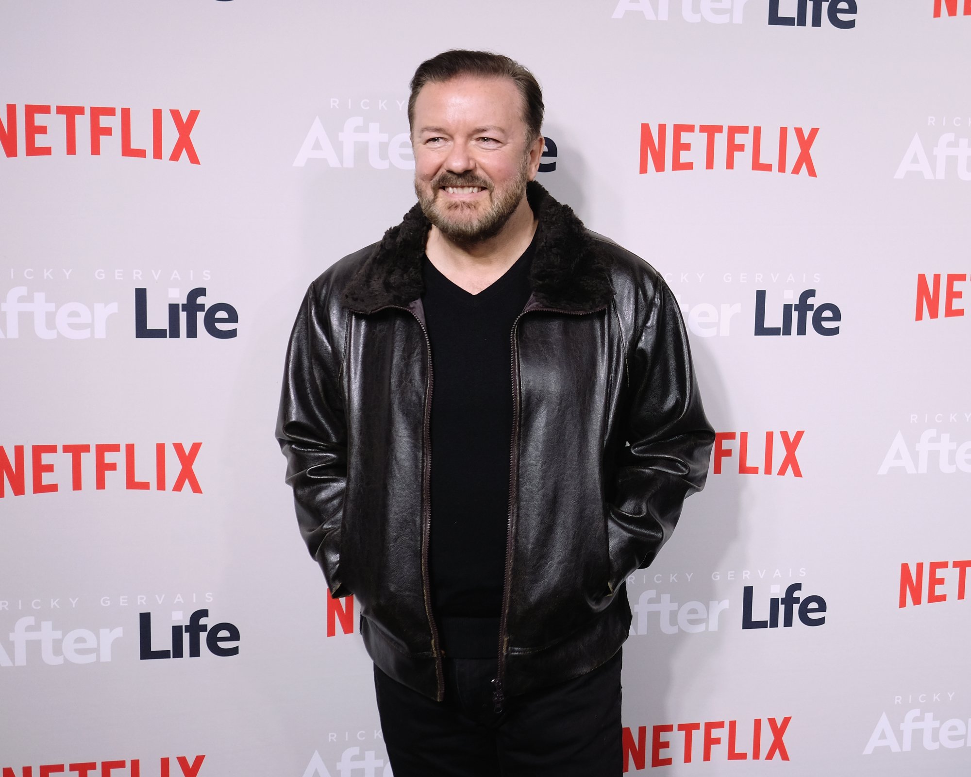 Ricky Gervais, who formulates his comedy from Twitter. He's wearing a leather jacket with his hands in his pockets in front of a step and repeat.