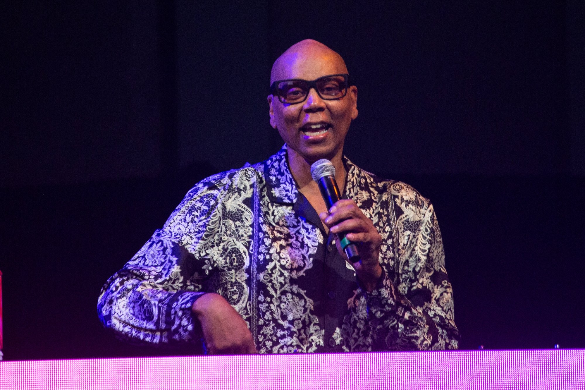 RuPaul, who owns multiple cars, speaking into a microphone wearing a black and white collared shirt
