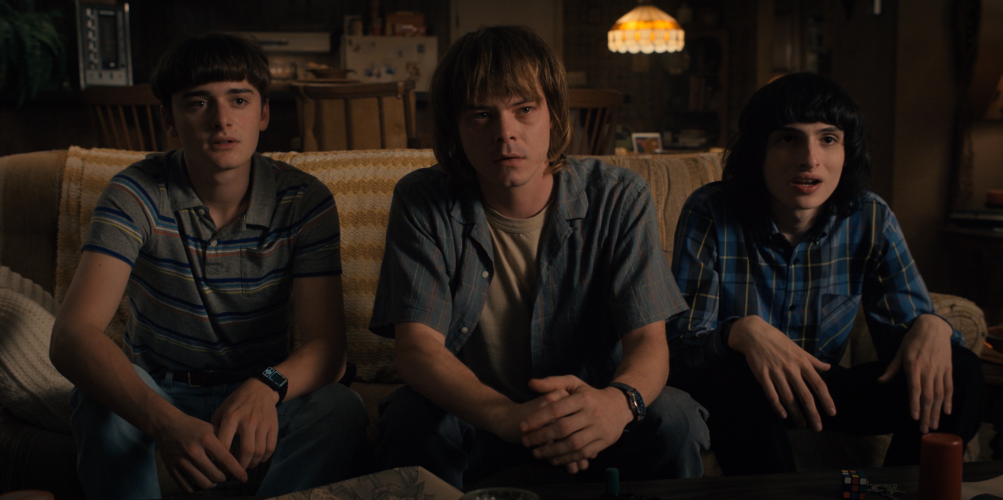 'Stranger Things 4' star Noah Schnapp, as Will Byers, sits on a couch next to Charlie Heaton and Finn Wolfhard in a production still.