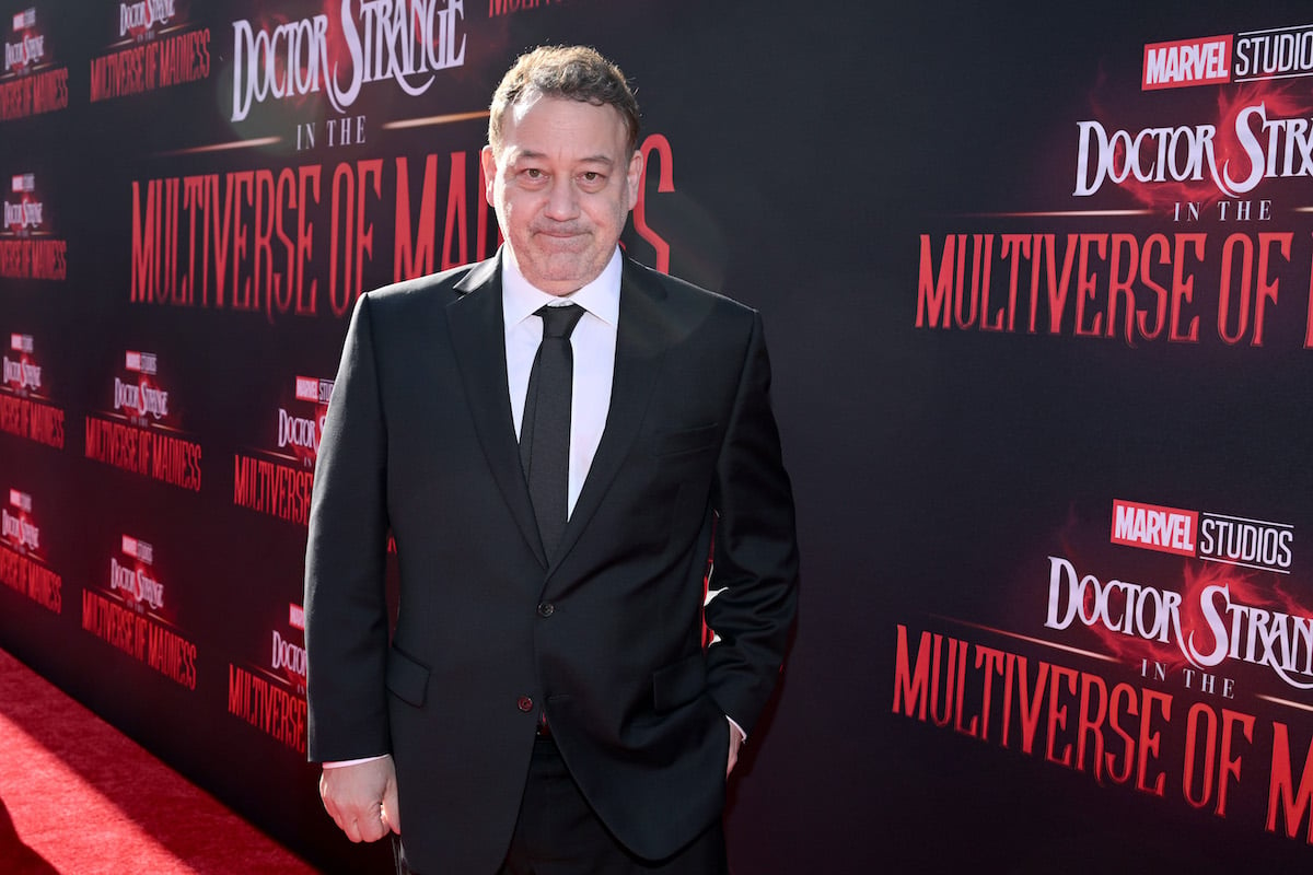 ‘Doctor Strange in the Multiverse of Madness’ director Sam Raimi wears a black suit and poses in front of the film’s logo